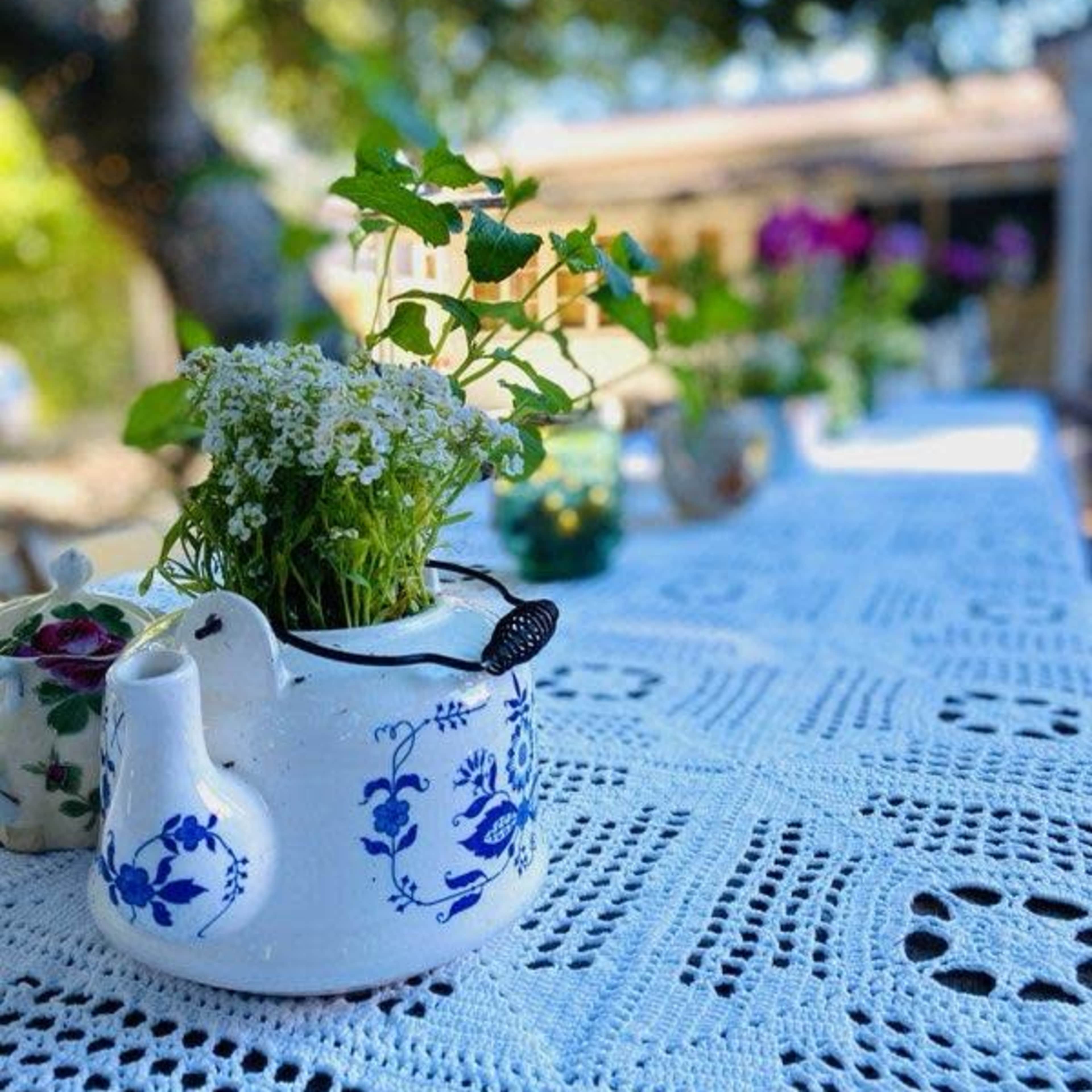 A teapot filled with flowers sits on a white lace tablecloth under trees in a garden setting.