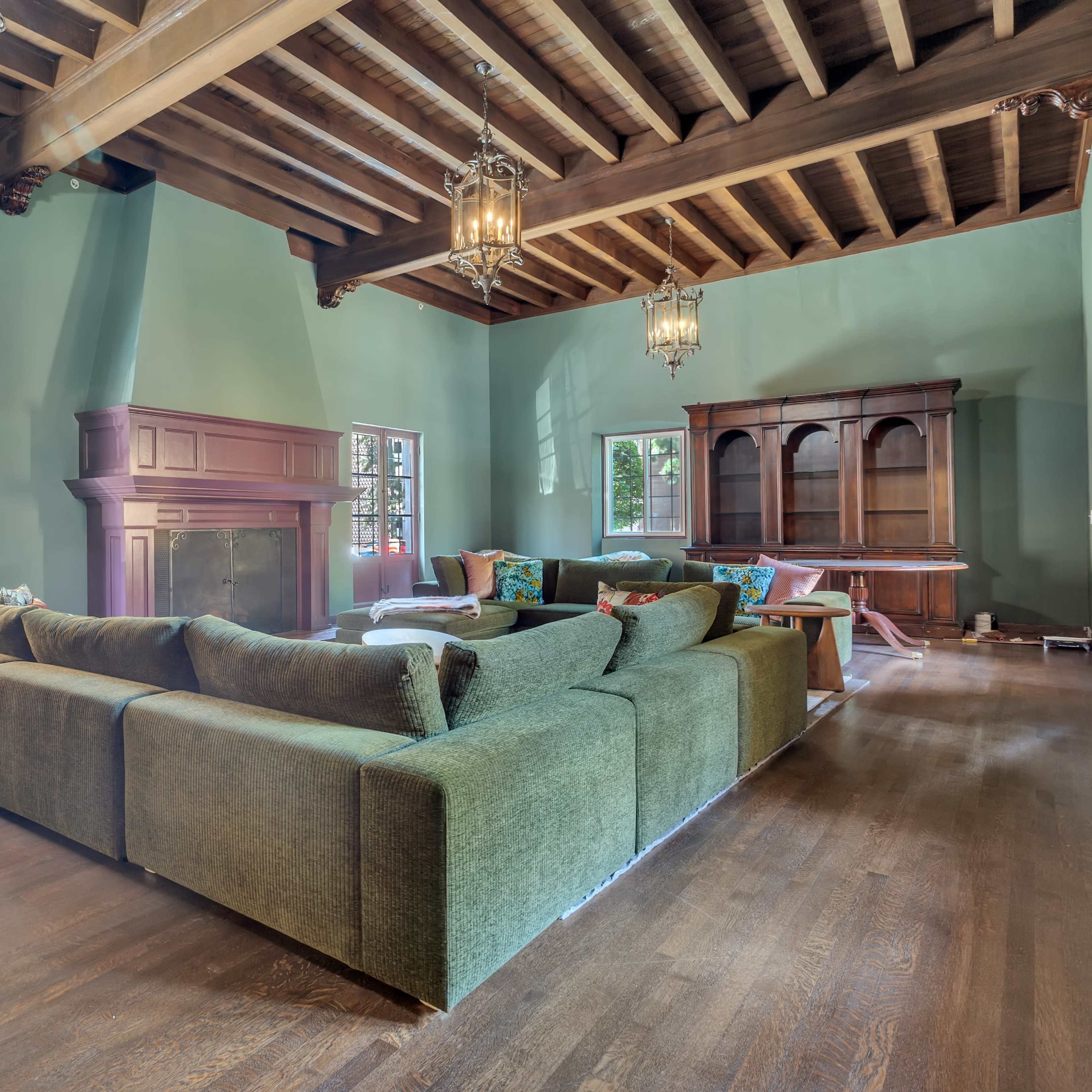 A spacious living room featuring a large green sectional sofa, wooden beams on the ceiling, and a fireplace with adjacent windows.