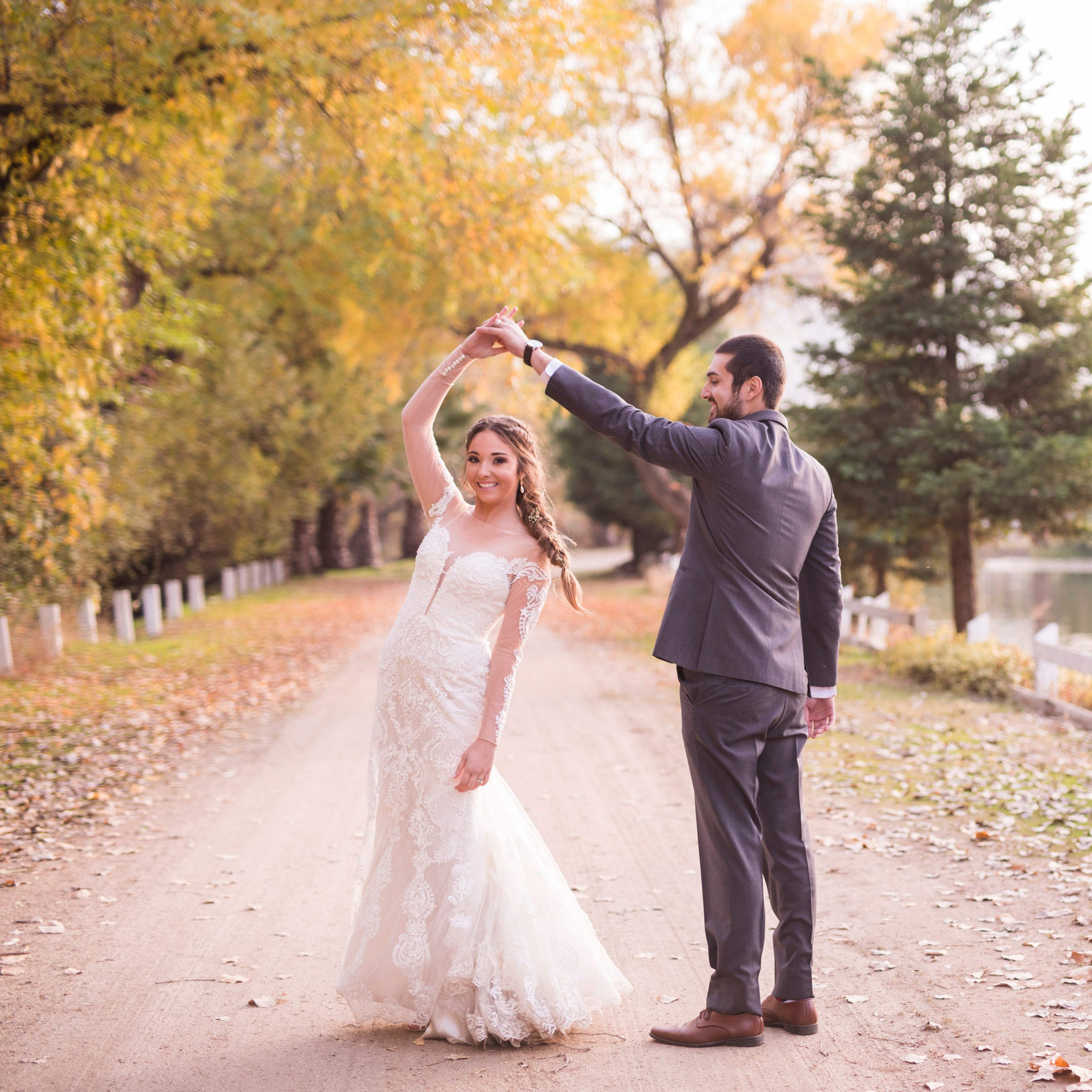 A couple in wedding attire dances along a tree-lined path covered in fall leaves, with a calm body of water visible nearby.