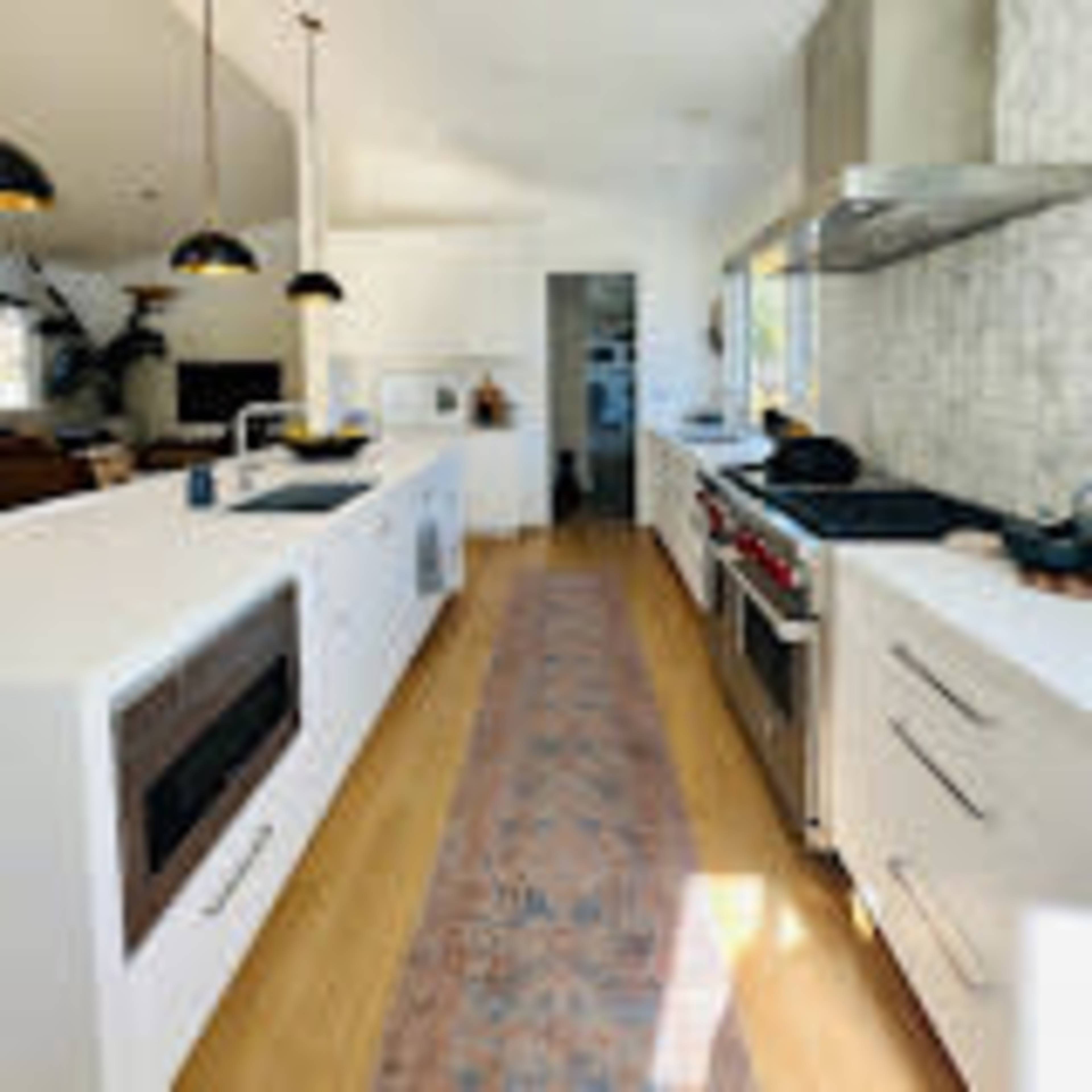 A modern kitchen features white cabinetry, stainless steel appliances, and a patterned rug running along the wood floor.