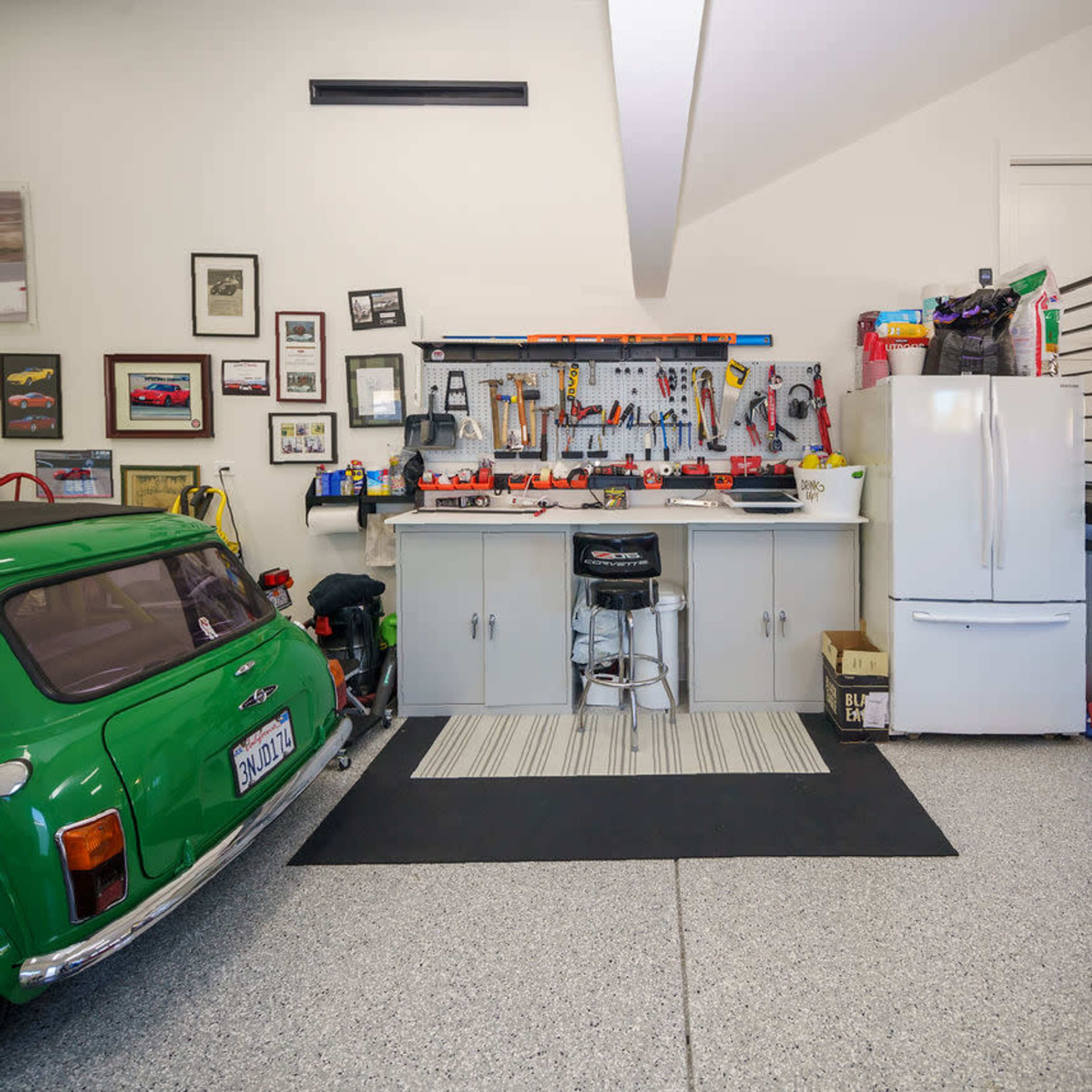 A garage featuring a green car, a workbench equipped with tools, a refrigerator, and bicycles parked on one side.