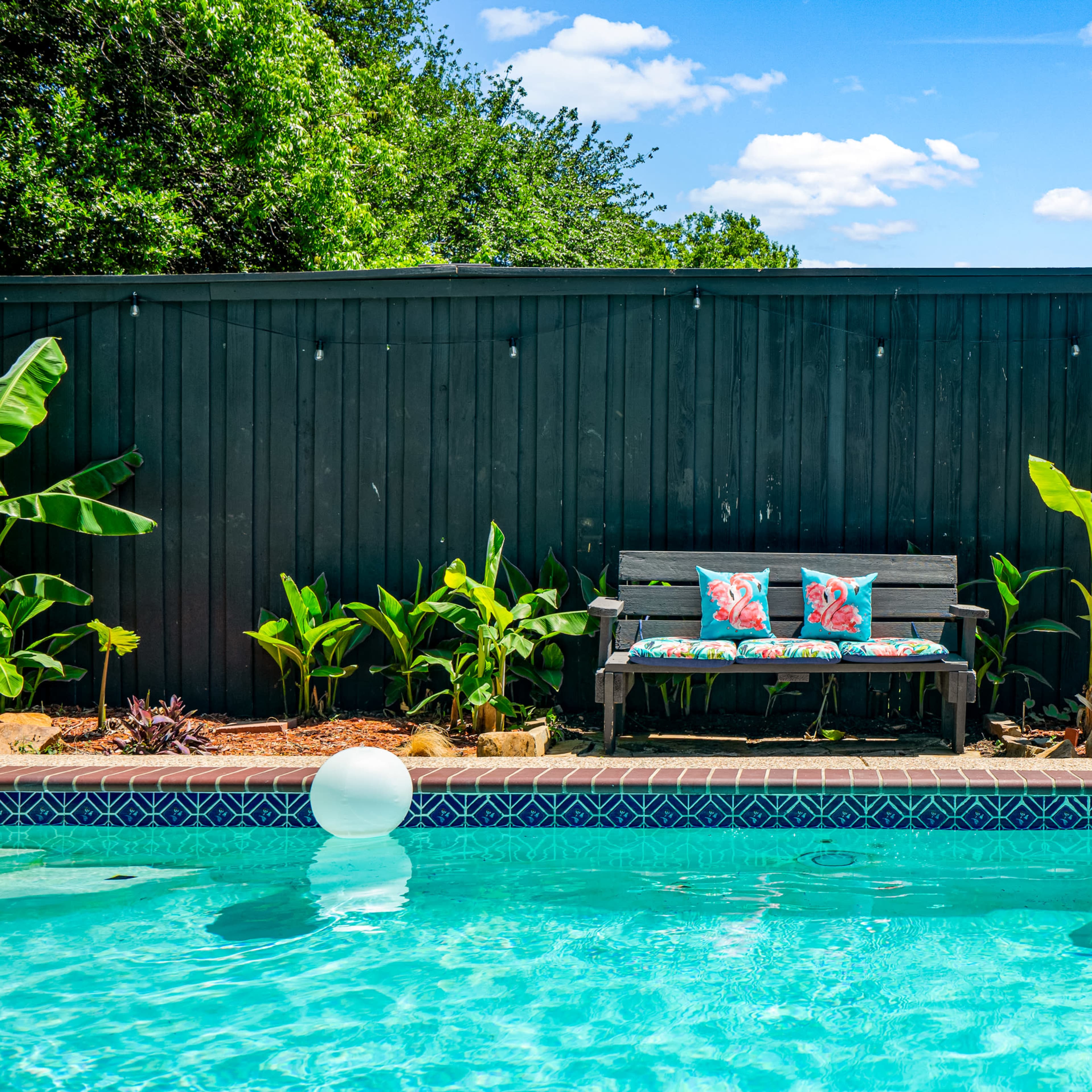 A wooden bench with colorful cushions is positioned beside a swimming pool, surrounded by greenery and inflatable floaties.