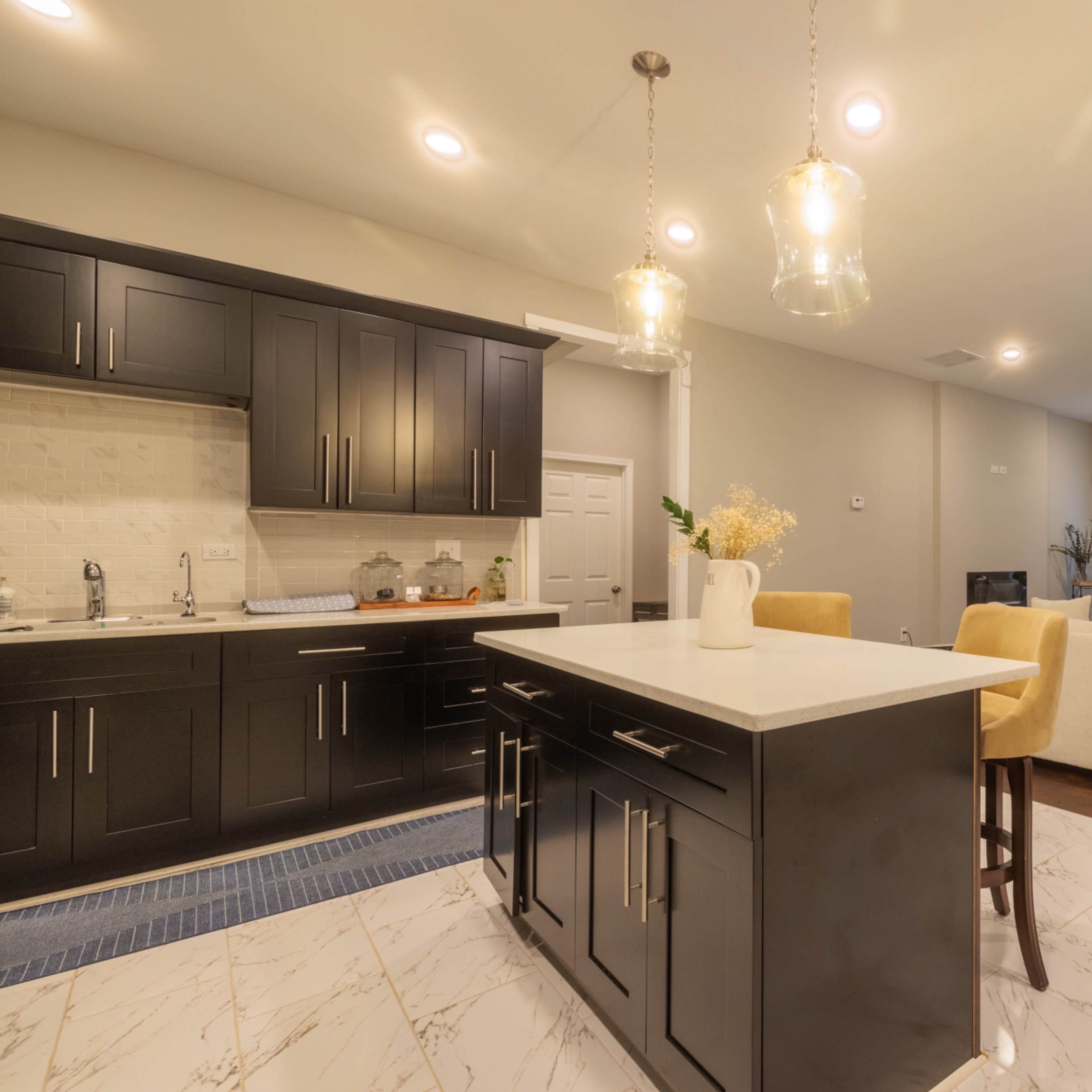 A modern kitchen with dark cabinetry, a central island with bar seating, and an adjoining living area furnished with a cream-colored sofa.