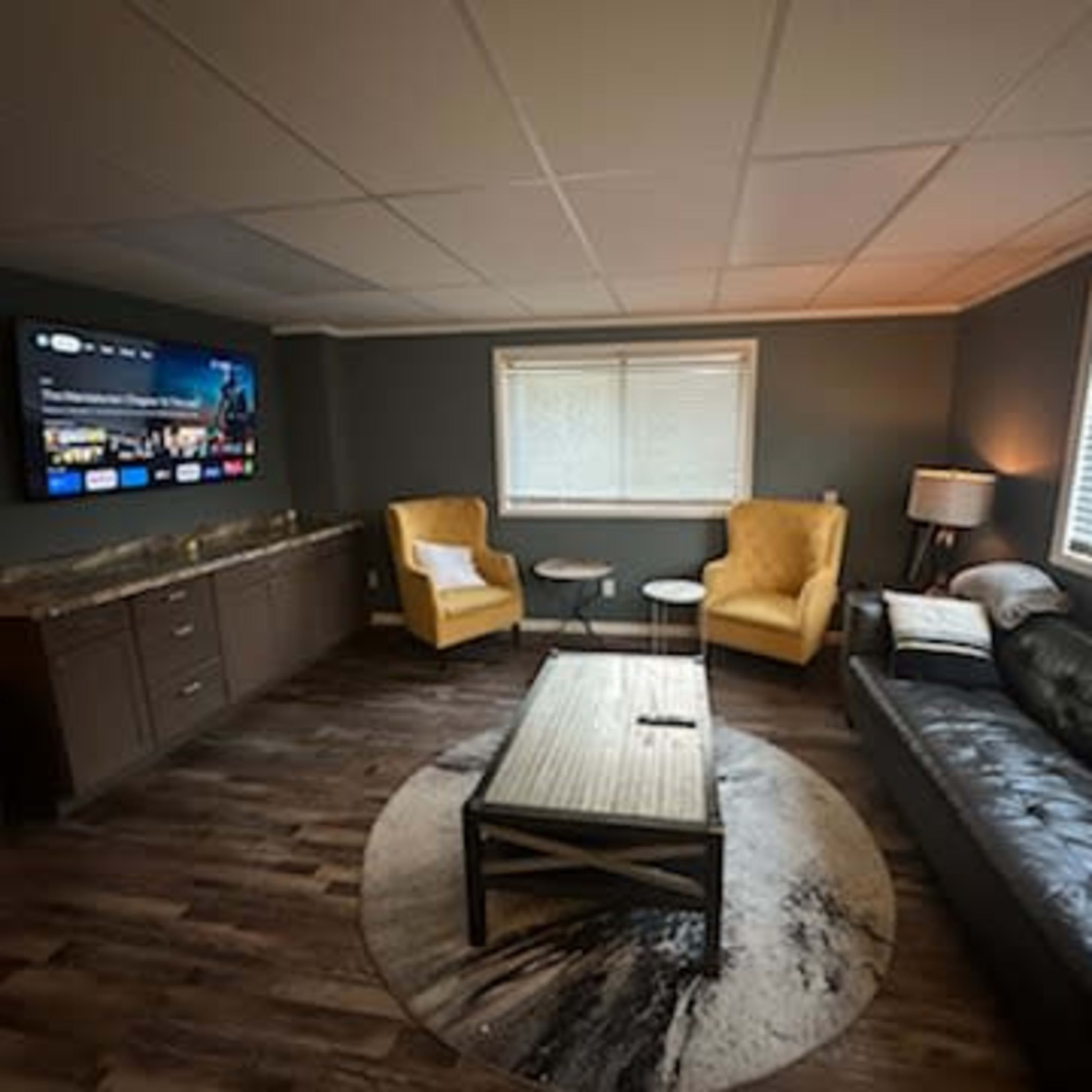 A modern living room with a black leather sofa, two yellow armchairs, a round rug, a coffee table, and a large wall-mounted TV.