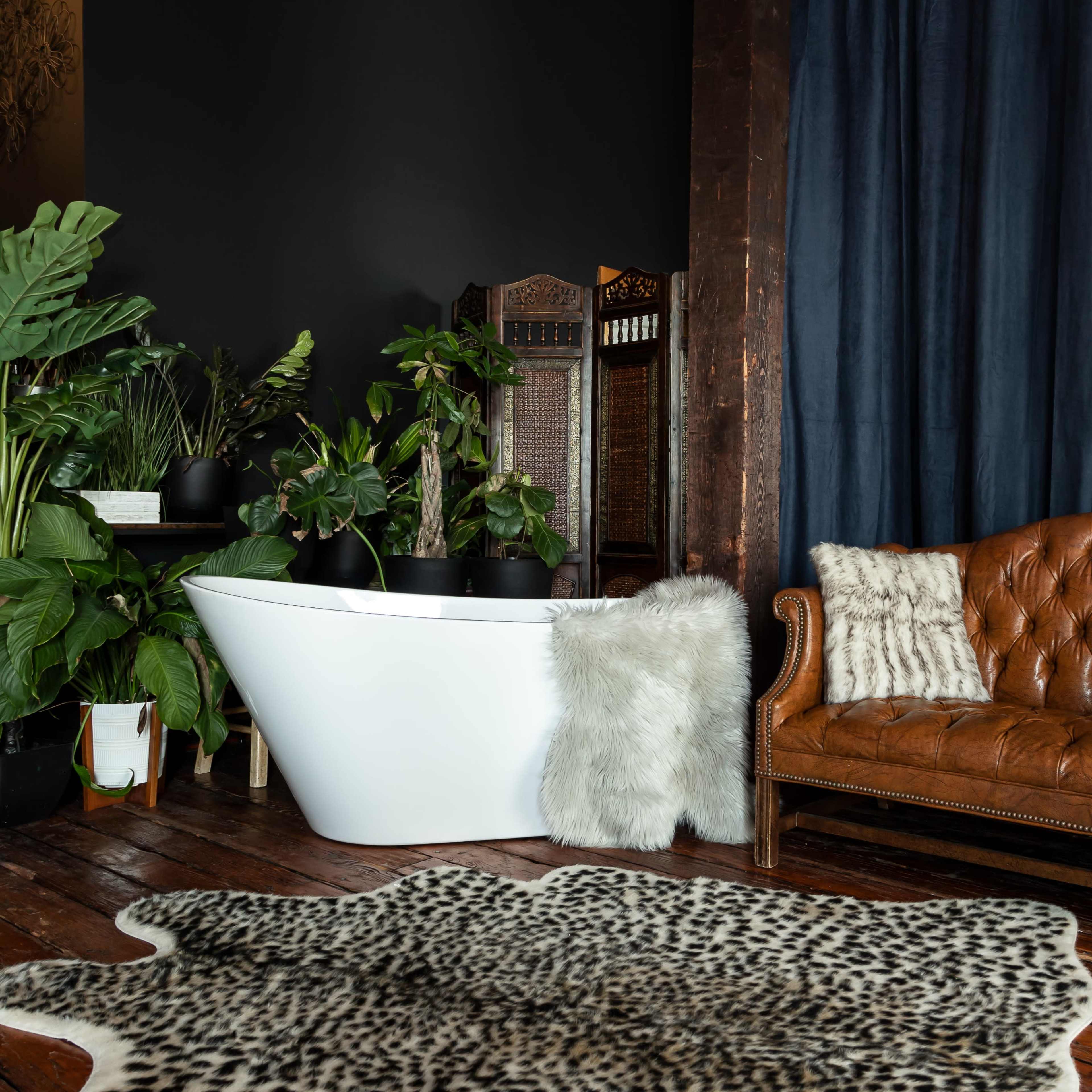 A stylish bathroom area with a freestanding white bathtub, a brown leather sofa, various indoor plants, and a patterned area rug on wooden flooring.