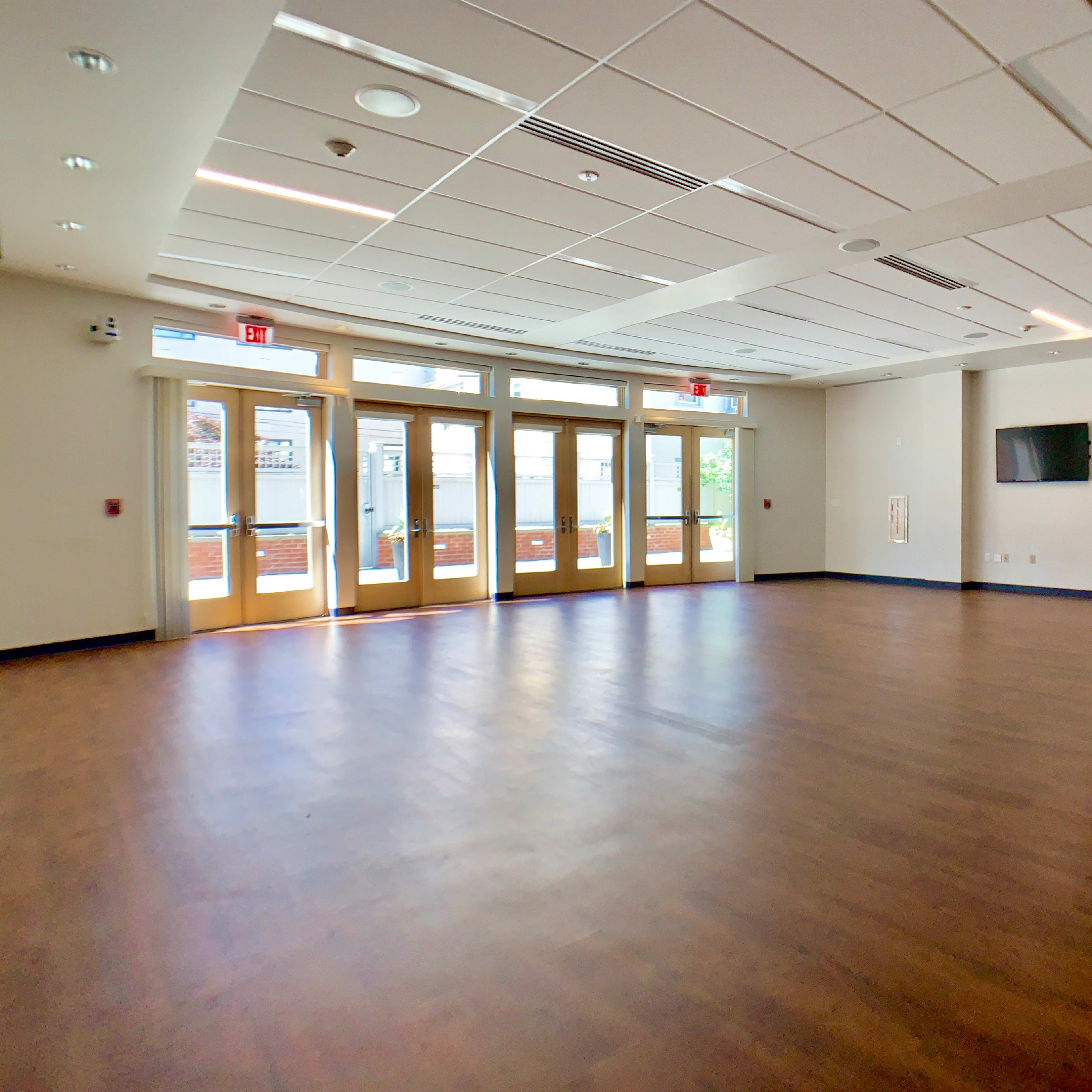 A spacious, empty room with wood flooring, large windows, and double doors leading outside.