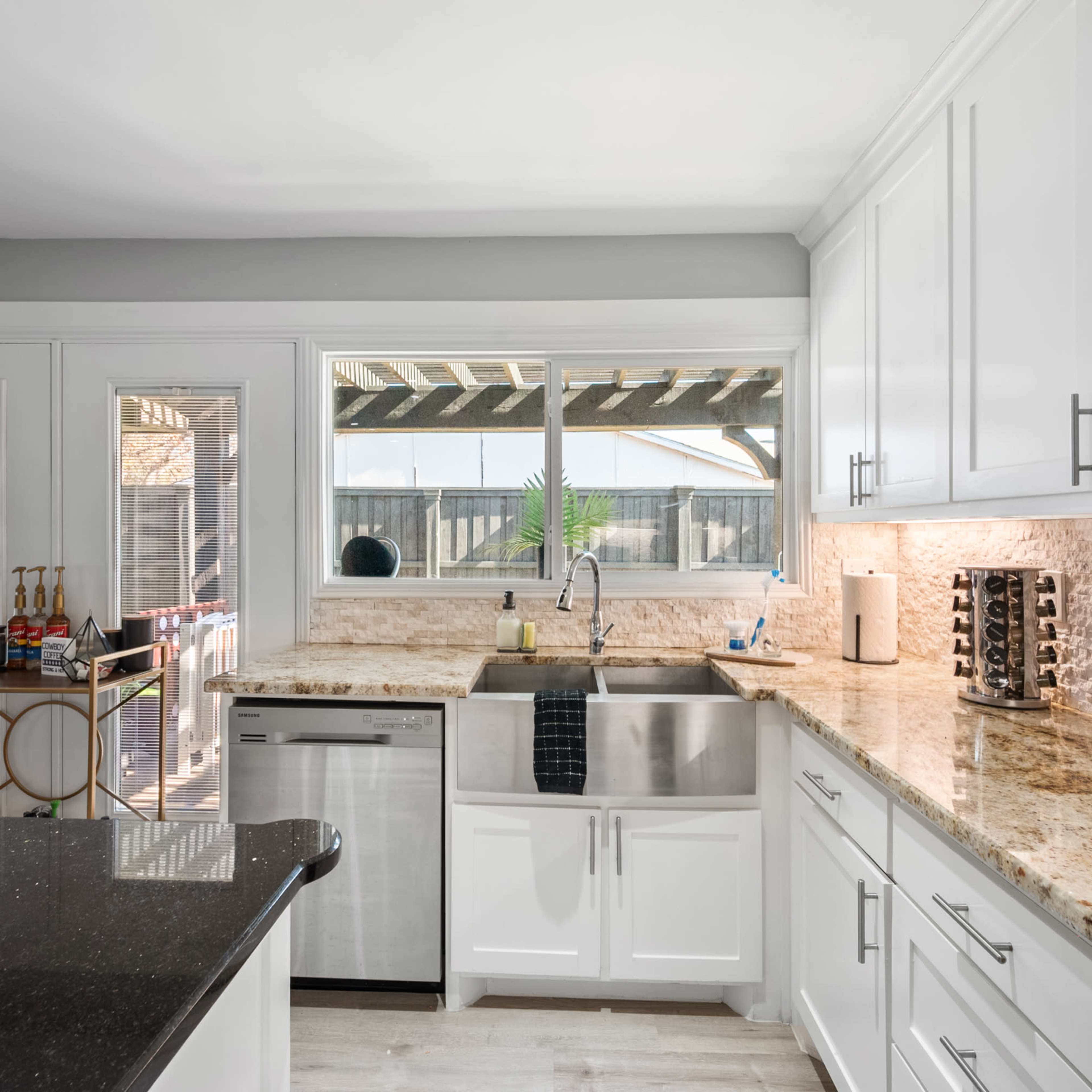 A modern kitchen features white cabinetry, a large sink beneath a window, and a dark countertop with stainless steel appliances.