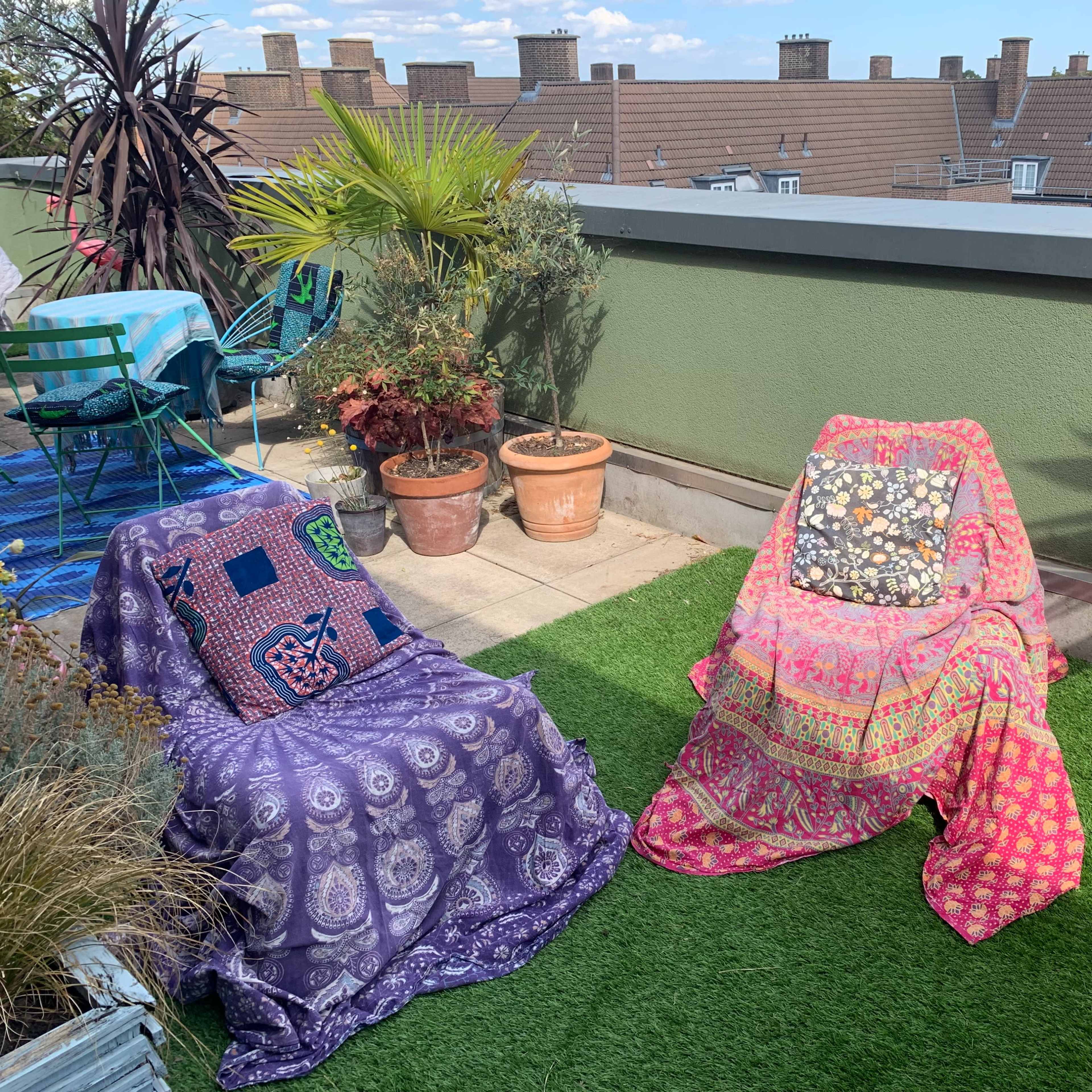 Two covered chairs adorned with vibrant textiles sit on a balcony featuring potted plants and a statue of Buddha.