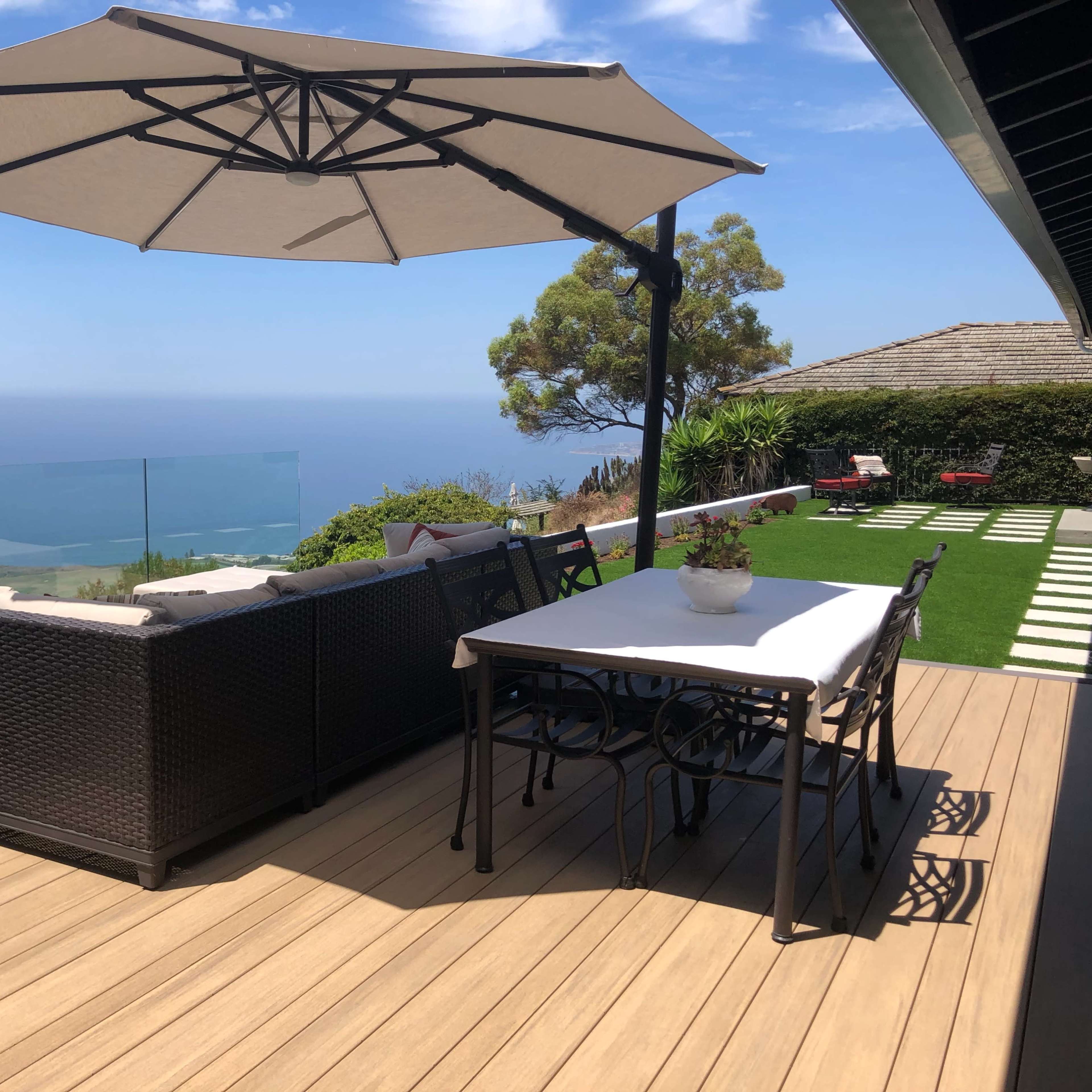 A patio with a sectional sofa, dining table, and large umbrella overlooks a scenic view of the ocean.