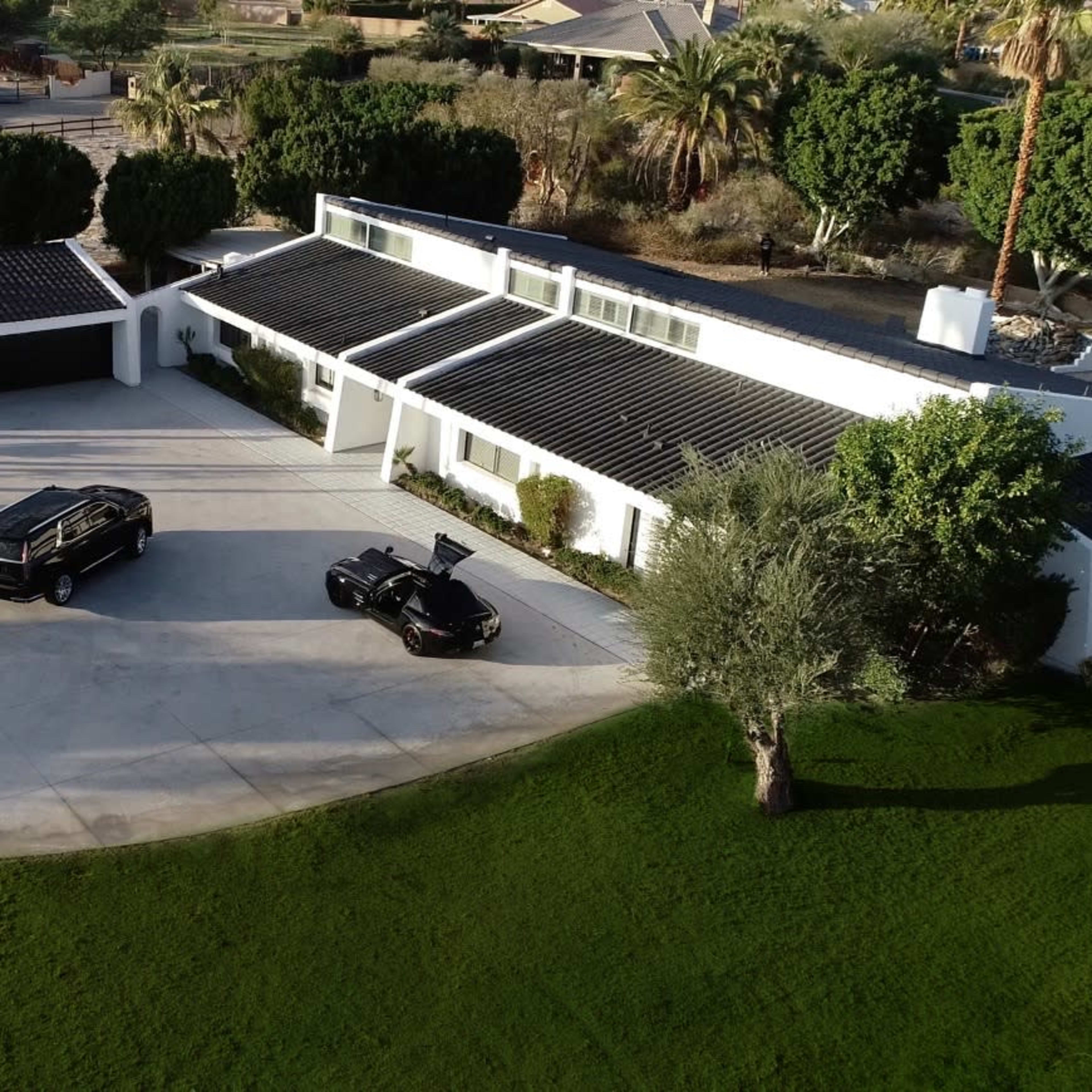 A modern white house with a black garage and two parked vehicles sits on a landscaped driveway surrounded by palm trees and green grass.