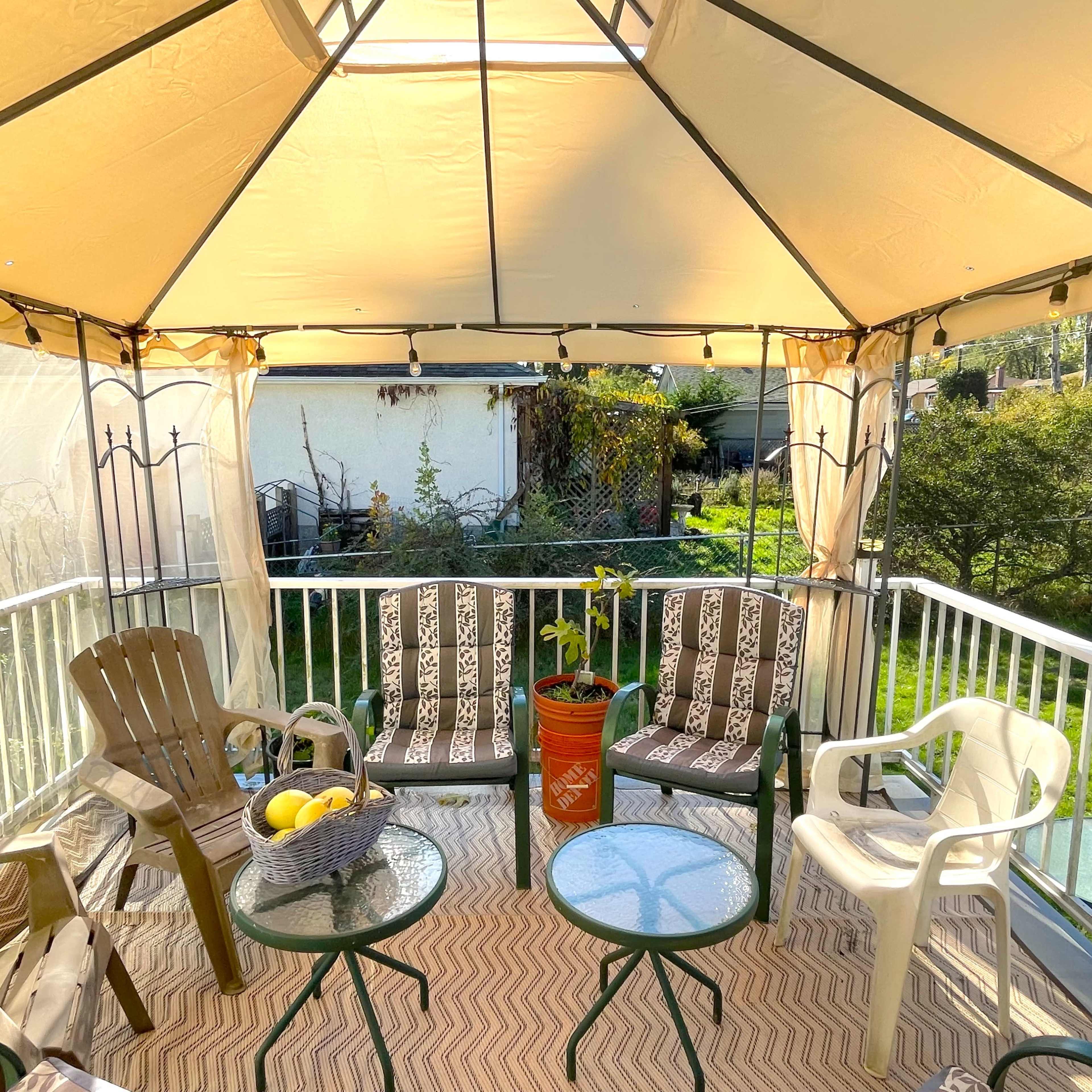 A covered patio area features several chairs, two small tables, and a potted plant, surrounded by greenery and a white railing.