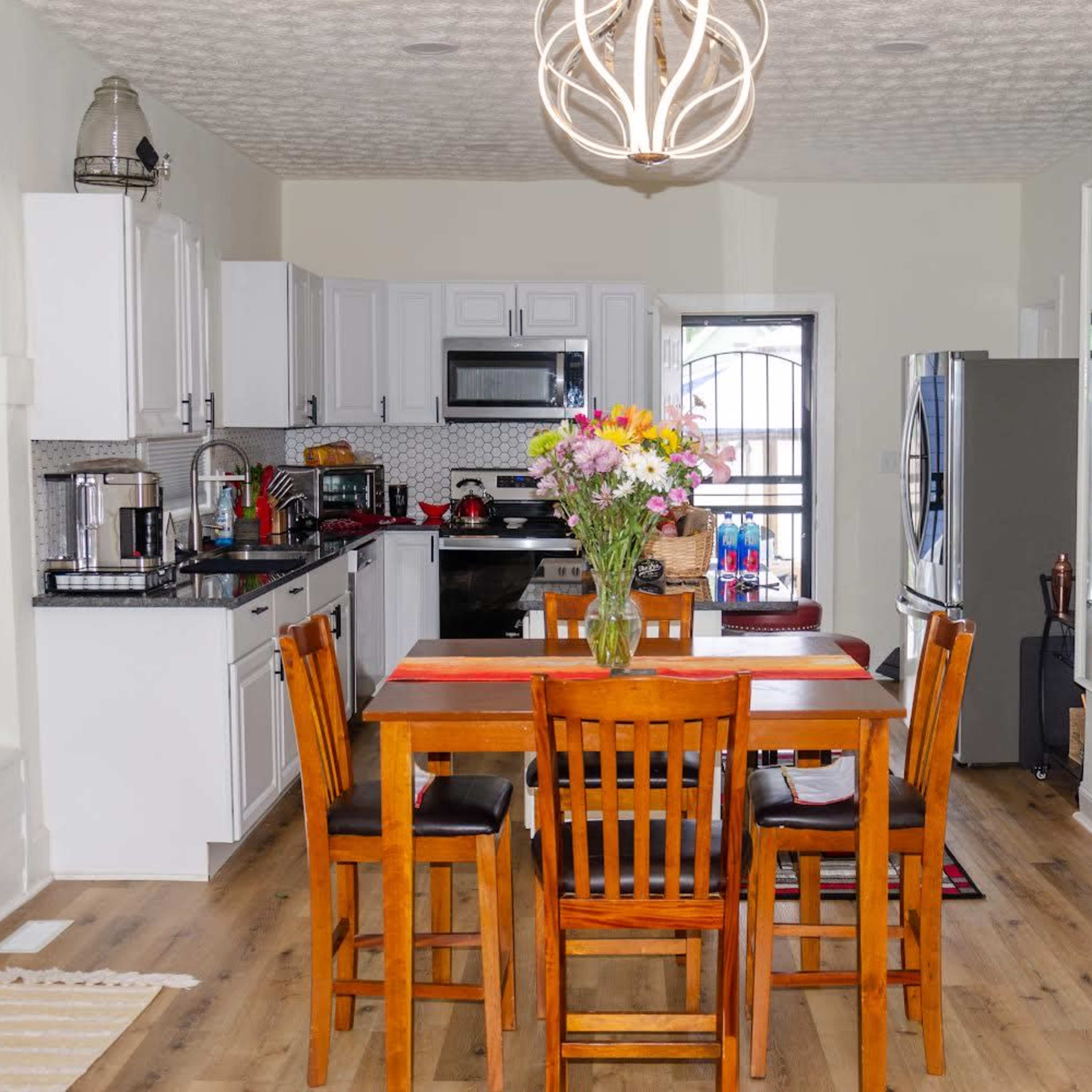 A modern kitchen features white cabinets, stainless steel appliances, a wooden dining table with four chairs, and a vase of flowers in the center.