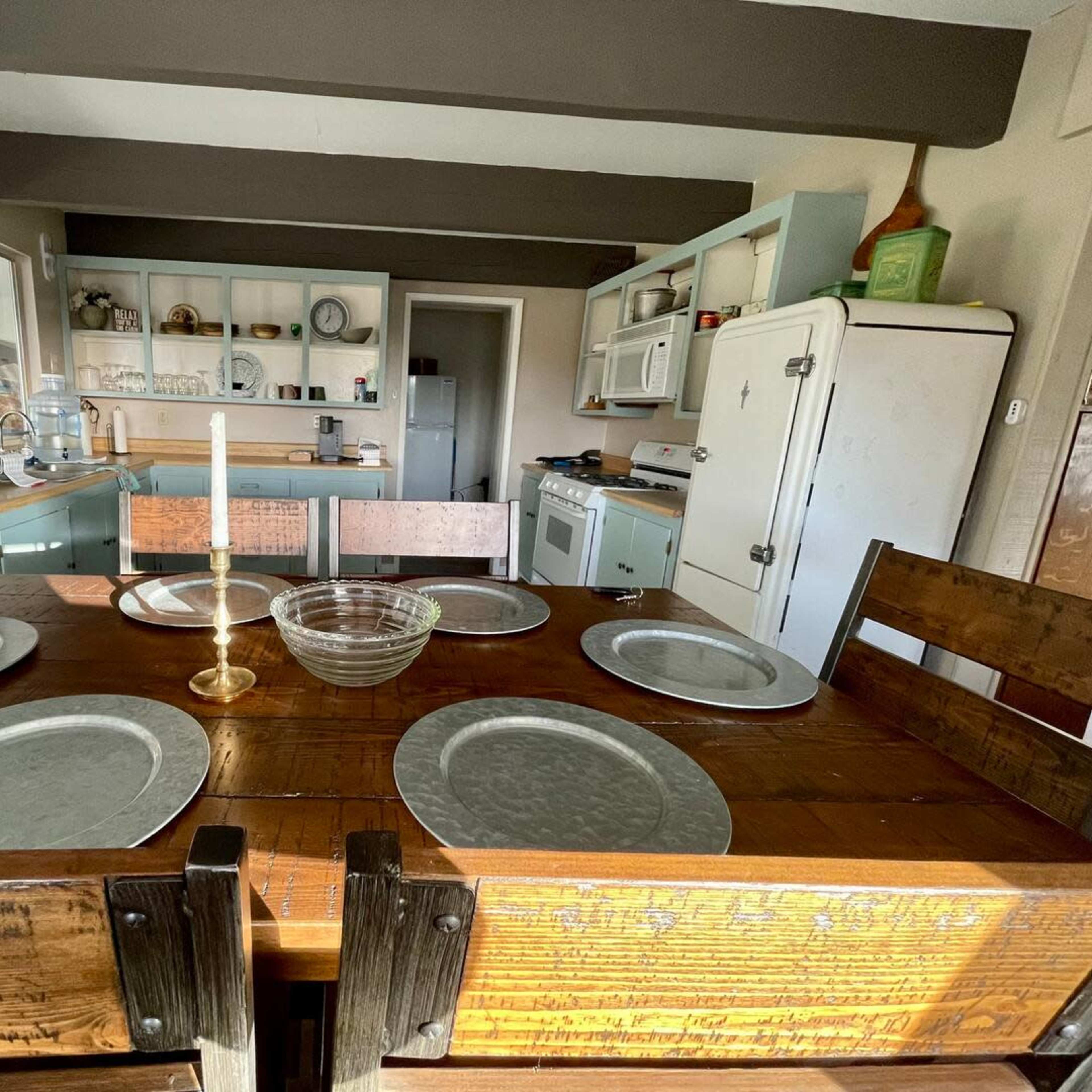 A rustic dining table set with round metal plates and a candle holder, situated in a kitchen with open shelving and a vintage refrigerator.