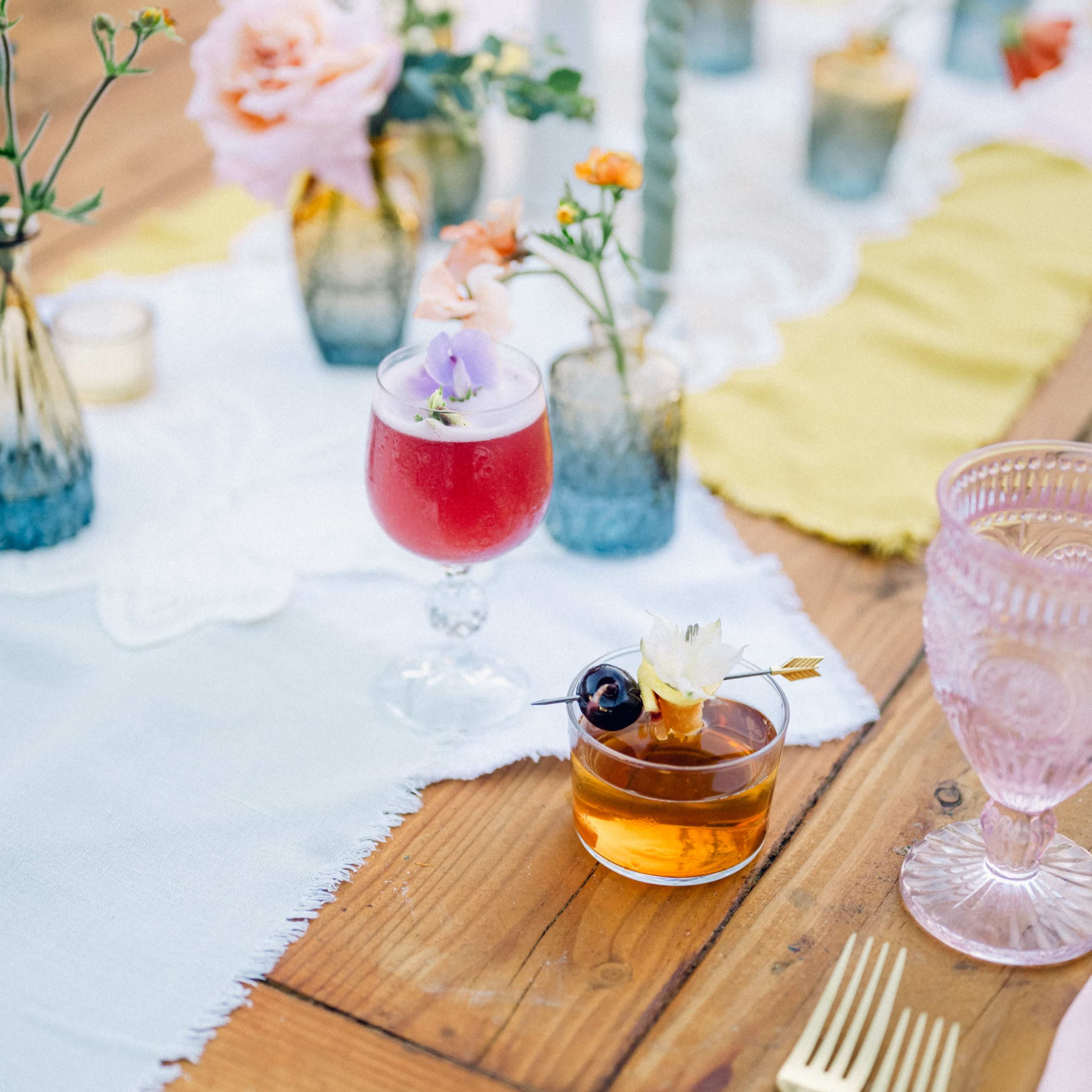 A wooden table is set with colorful drinks, floral arrangements in varying glass vases, and elegant tableware.
