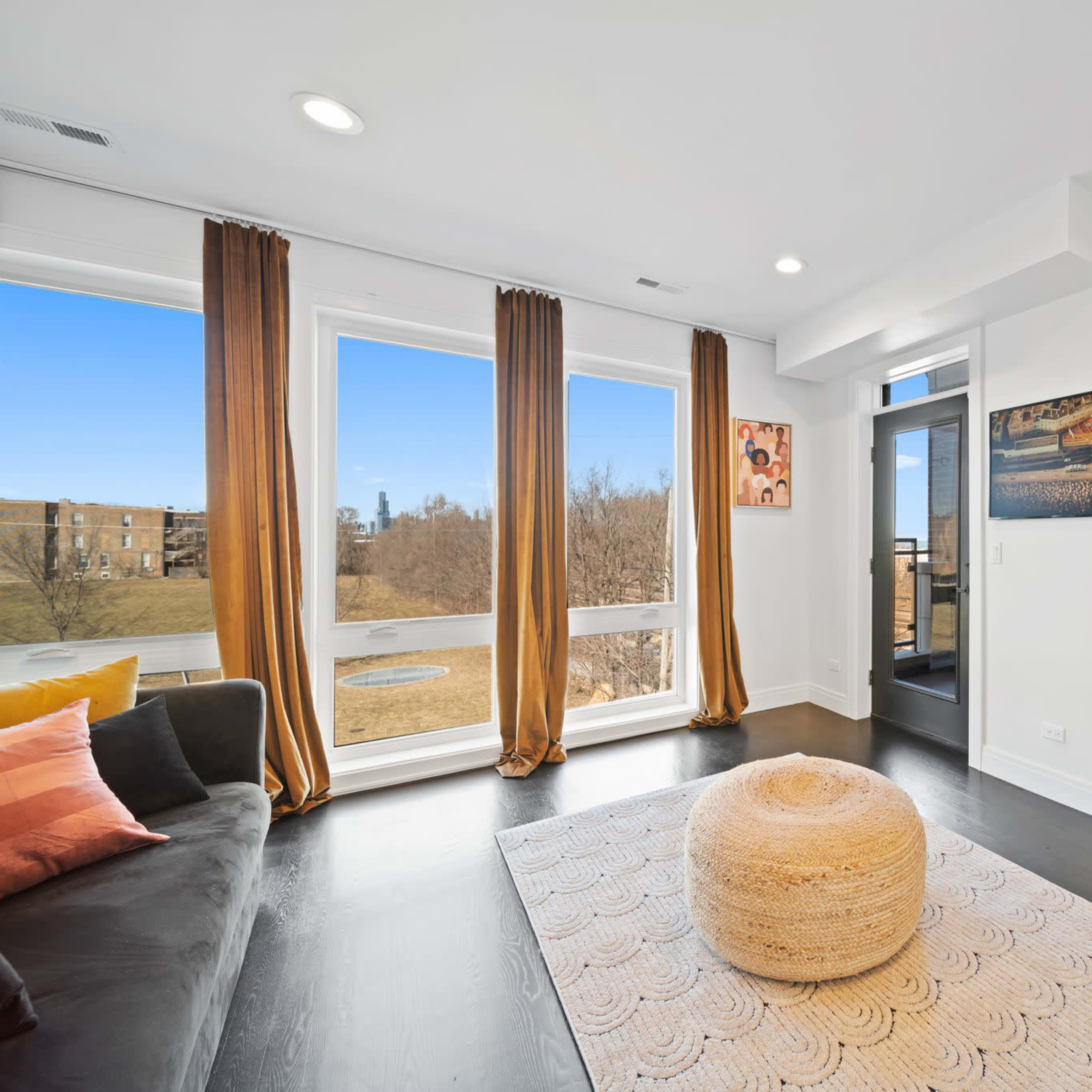 A modern living room features large windows with light curtains, a sofa, a circular ottoman, a decorative rug, and a view of a landscape outside.