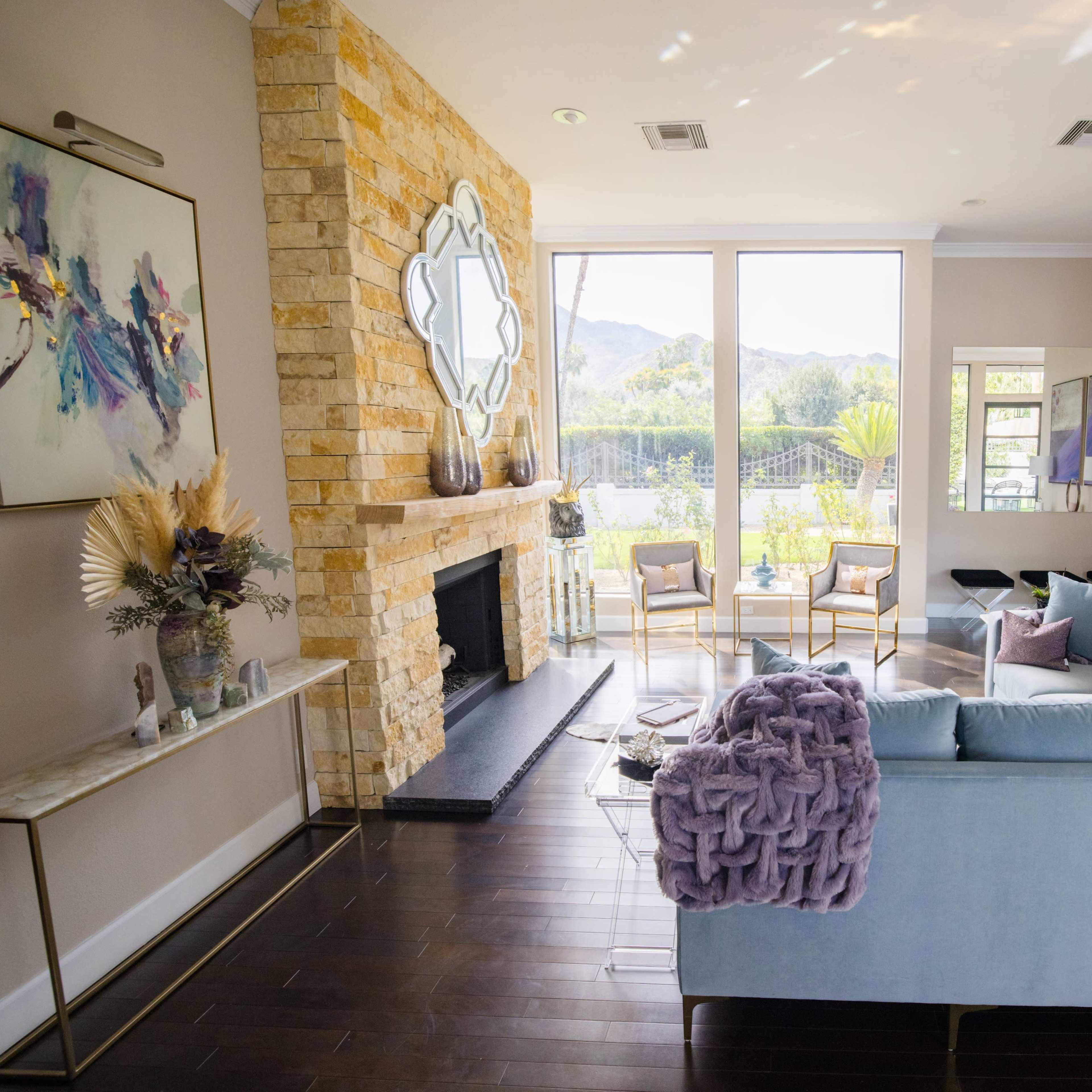 The room features a light-colored sofa with a textured throw, a stone fireplace, and large windows overlooking a patio and greenery.