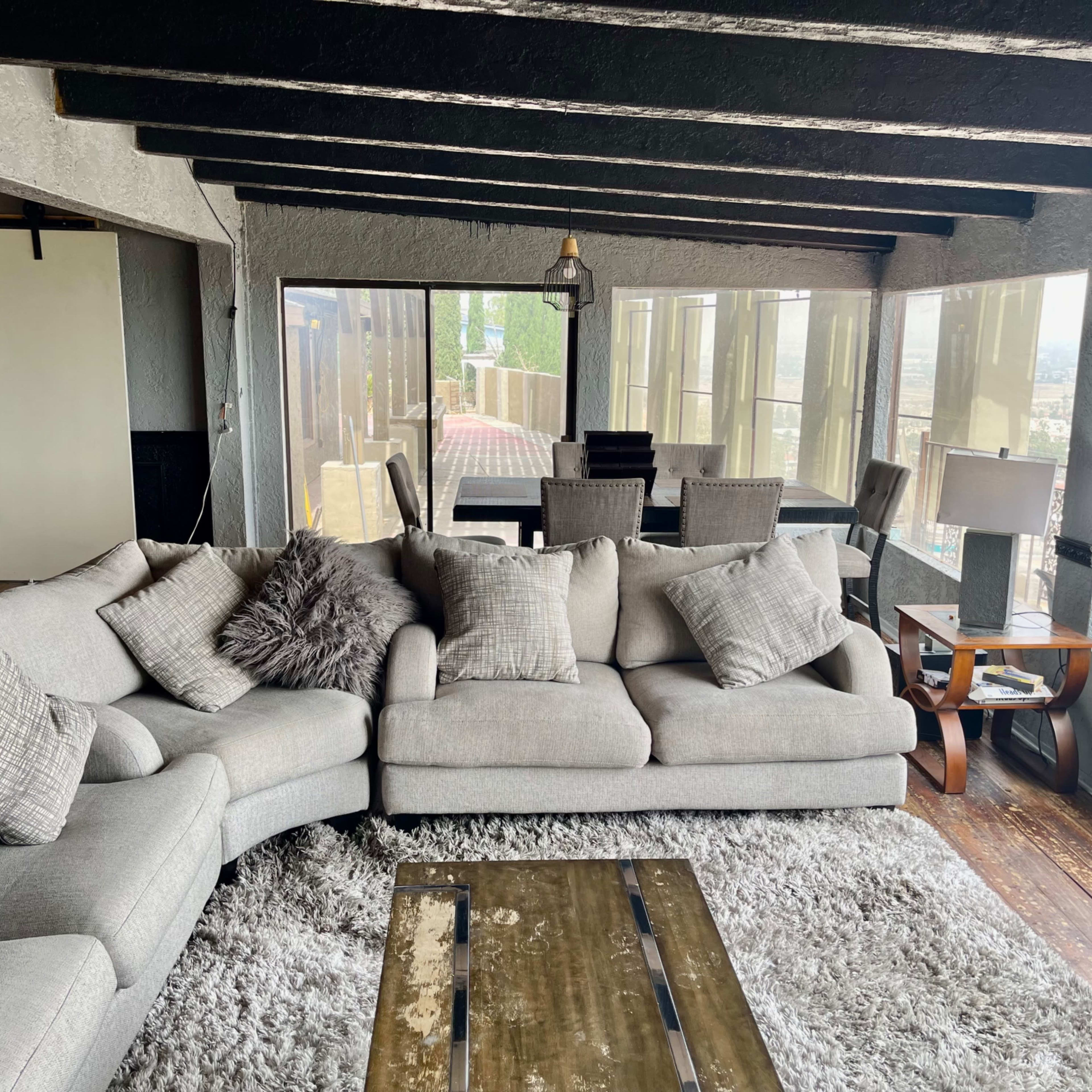 A light-colored sectional sofa with decorative pillows, a wooden coffee table, and large windows overlooking a scenic view, within a room that has exposed wooden beams and a cozy atmosphere.