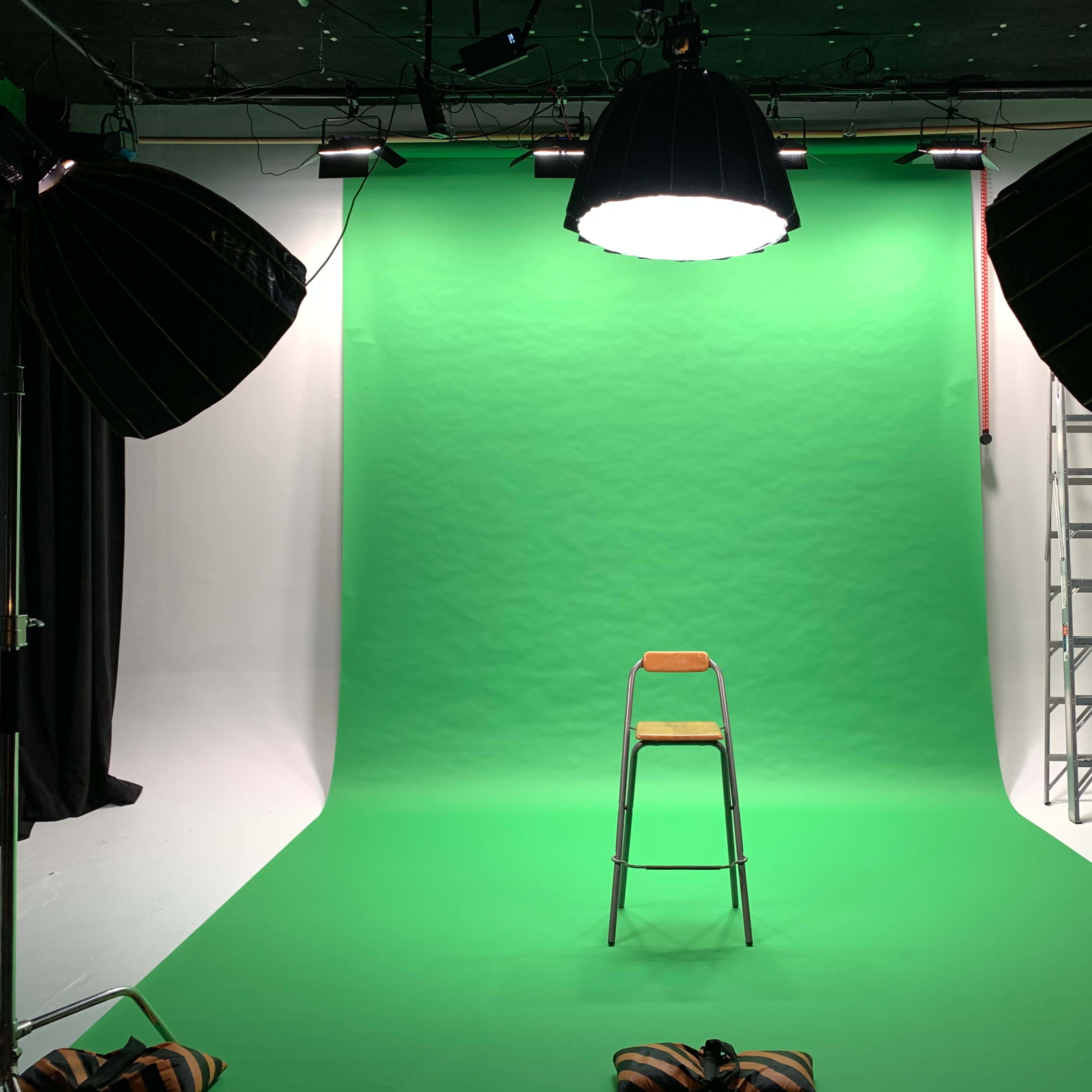 A studio setup features a green screen backdrop with a metal chair in the center, flanked by large studio lights and a ladder in the background.
