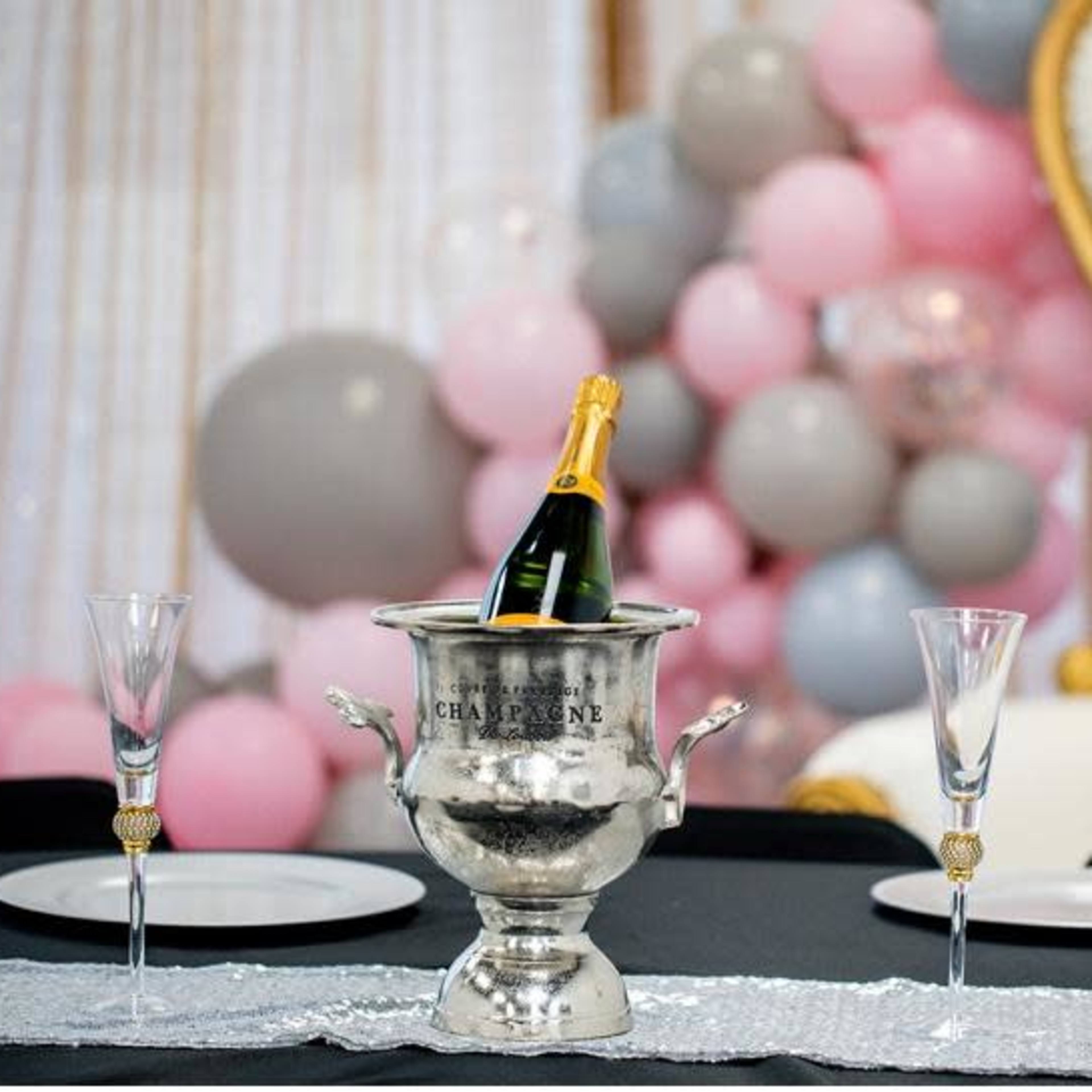 A silver champagne bucket with a bottle of champagne sits on a table with elegant glassware, while a backdrop of pink, gray, and white balloons adds decoration.