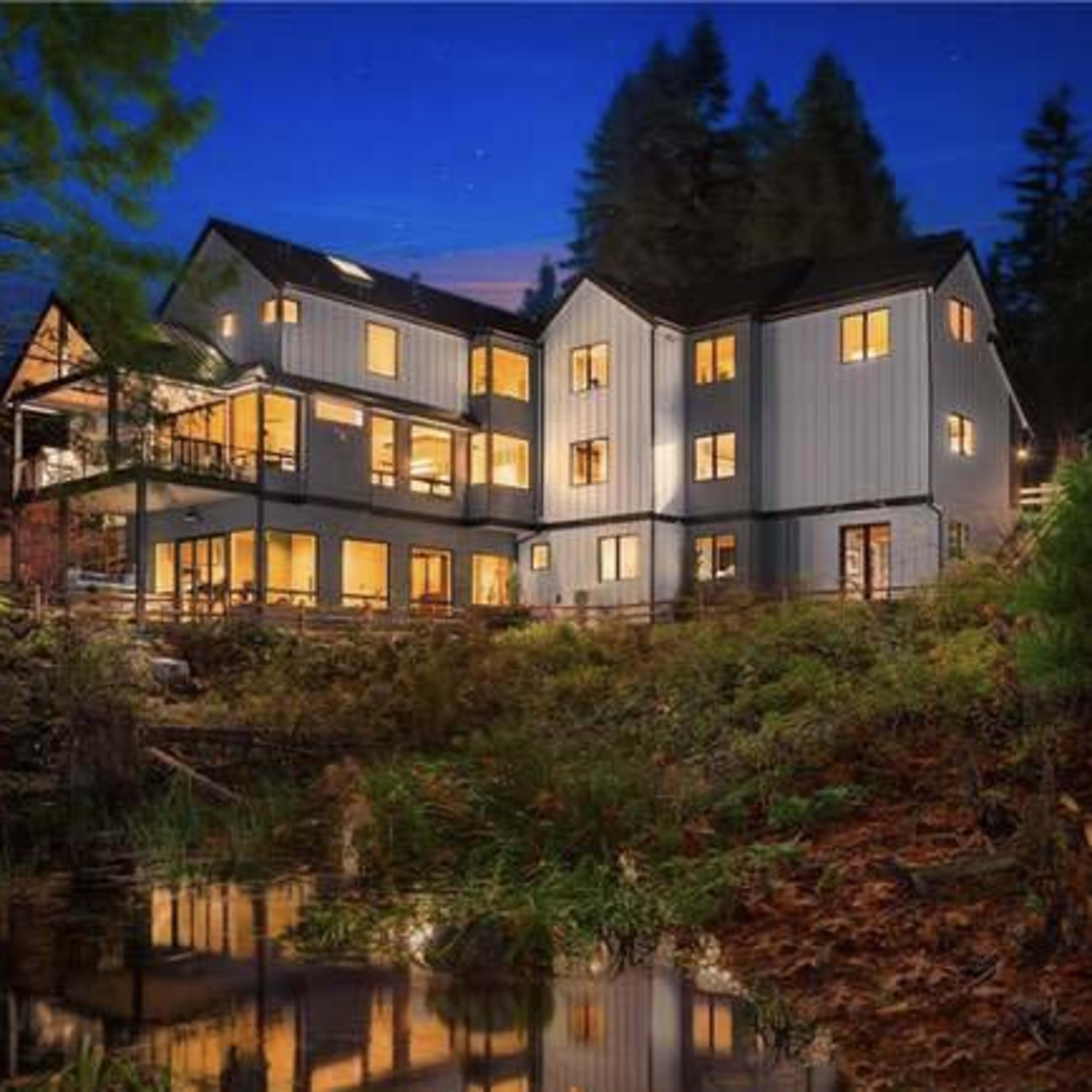 A modern multi-story house with large windows is situated by a calm body of water, surrounded by trees at dusk.
