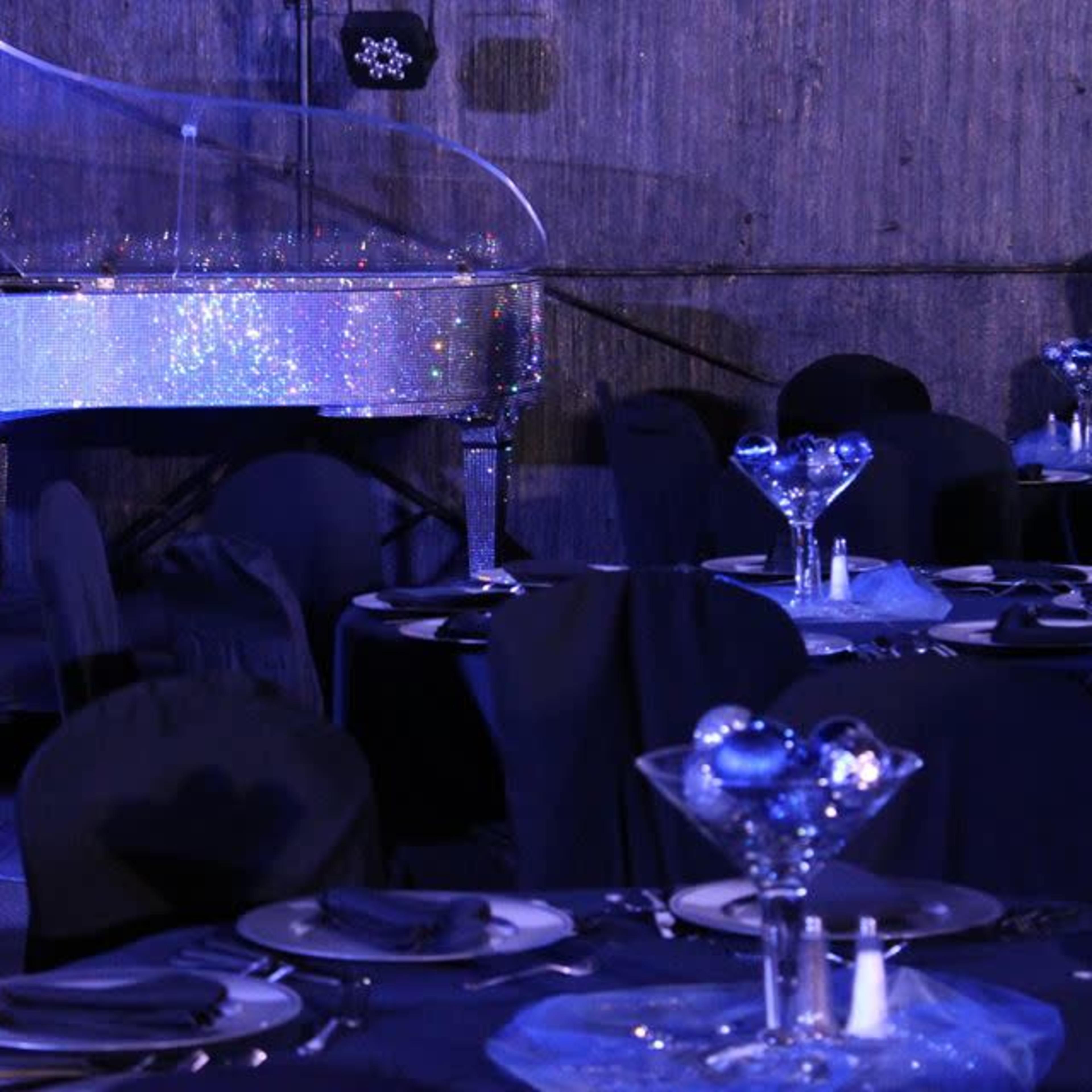 A sparkling clear grand piano is positioned beside elegantly set tables adorned with black covers and decorative glassware.