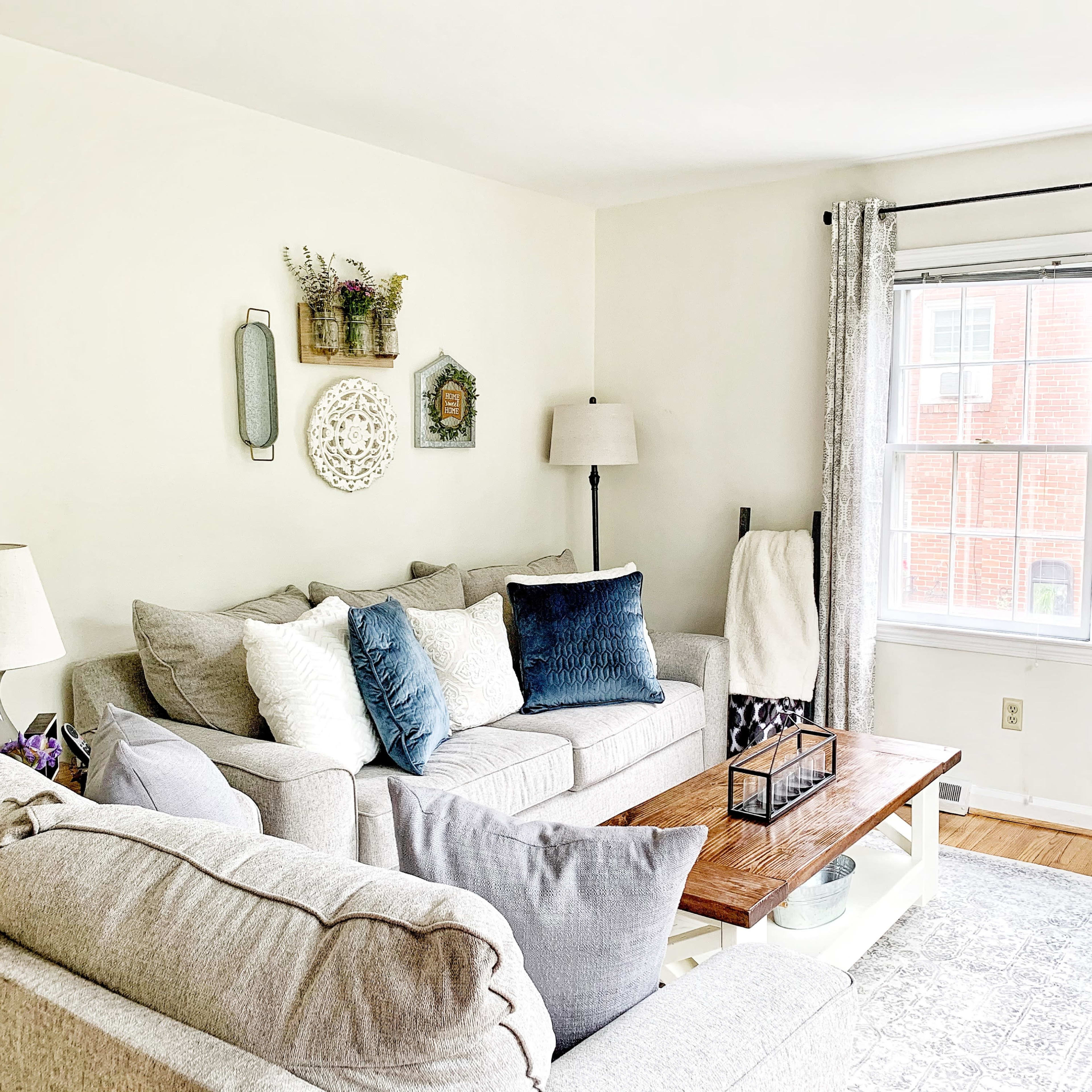 A cozy living room with light-colored sofas, a wooden coffee table, and decorative wall hangings near a window.