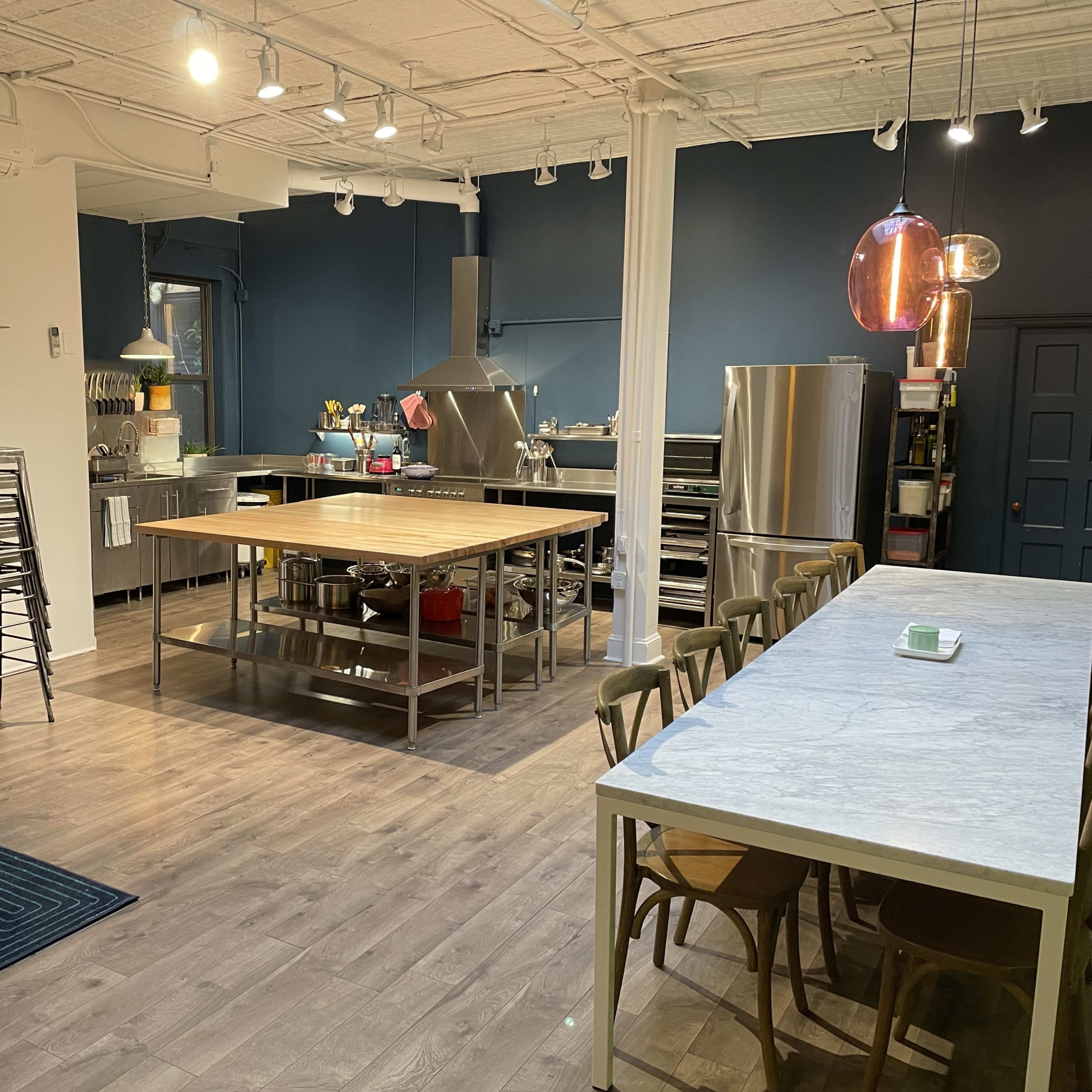 A modern kitchen and dining area features a large central table, stainless steel appliances, and a pink velvet sofa against the wall.