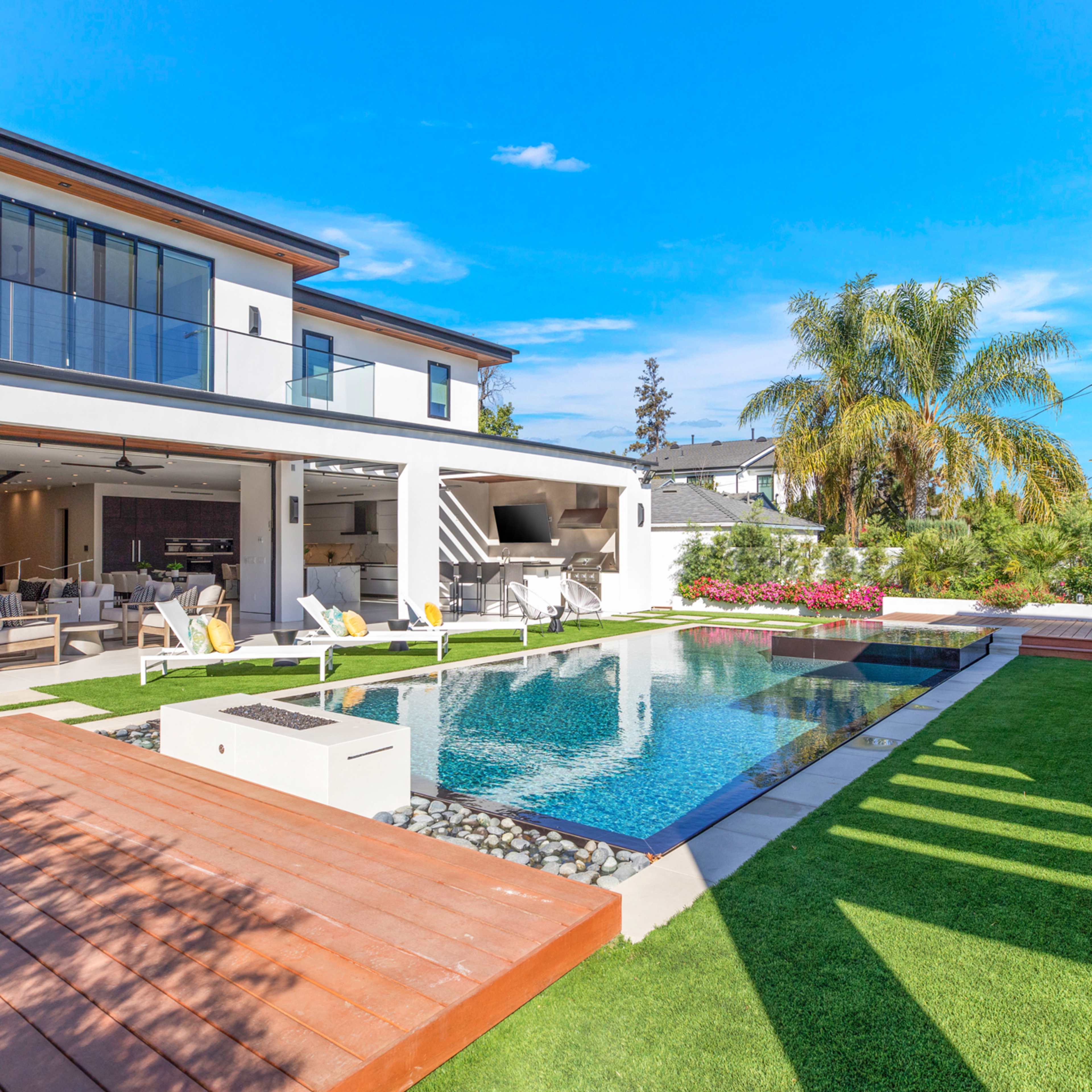 A modern residential backyard featuring a large pool, surrounding lounge chairs, and a well-designed outdoor space with landscaped greenery and a two-story house.