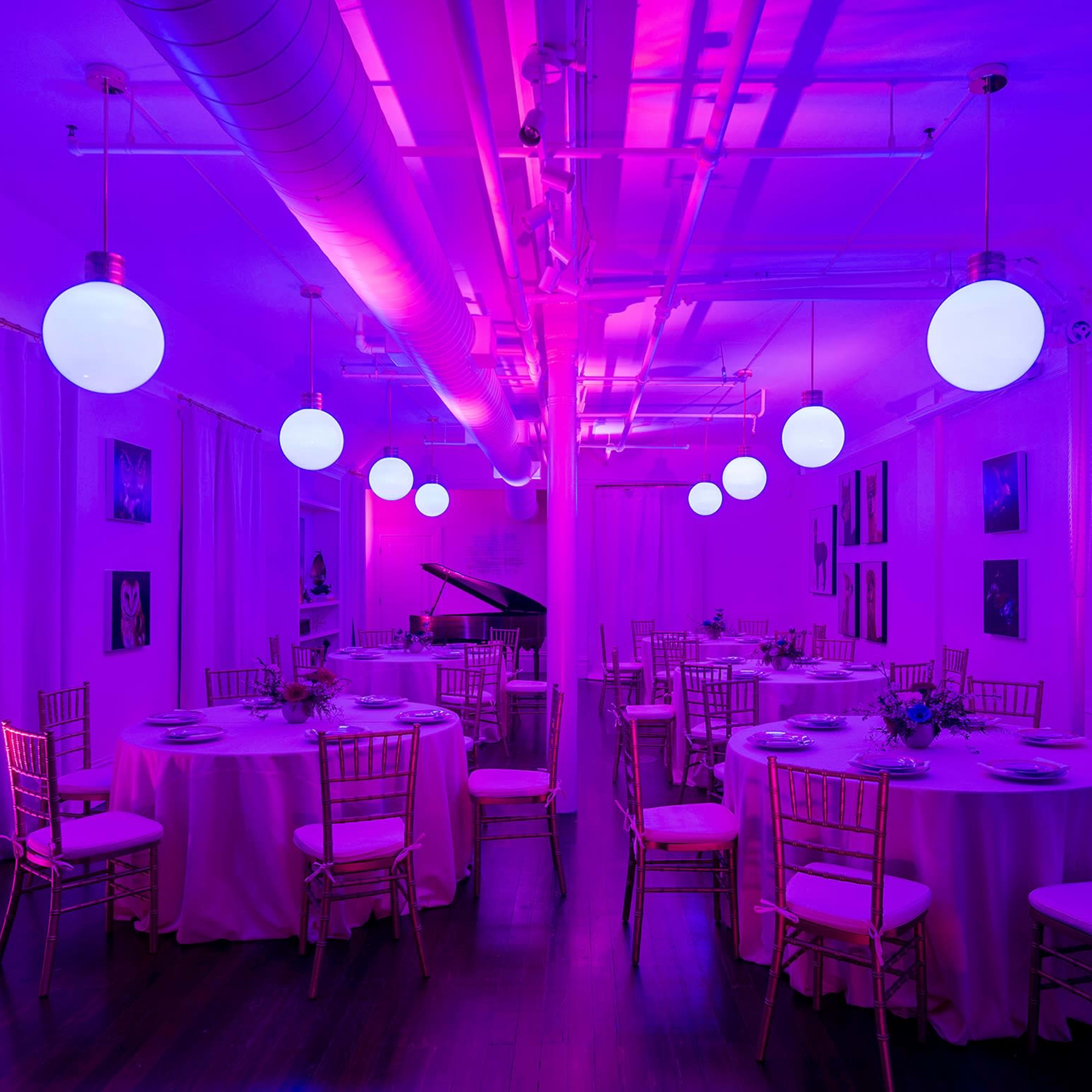 A decorated event space features circular tables with pink tablecloths, surrounded by gold chairs and illuminated by glowing purple lights.