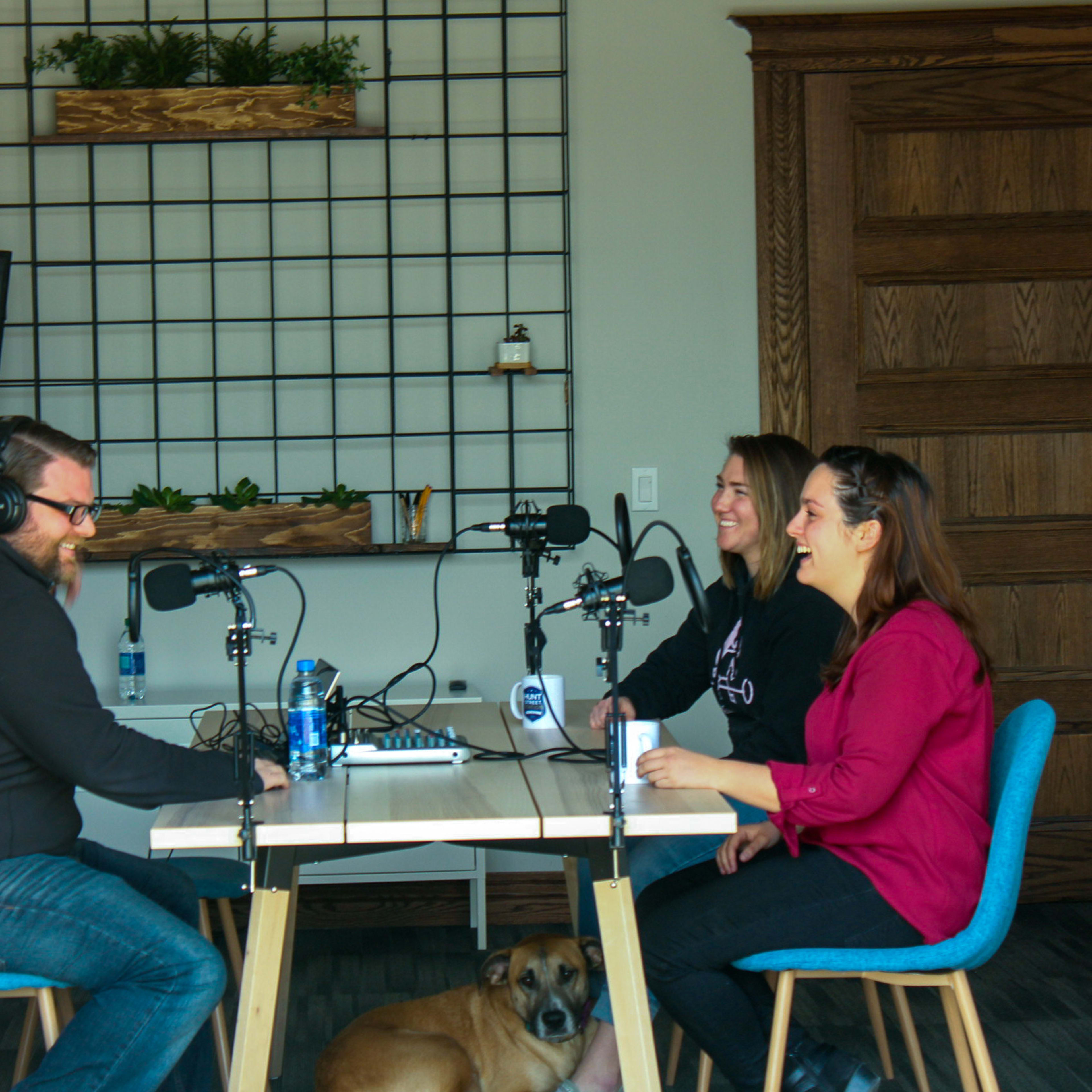 Three people sit at a table with microphones in a room featuring a grid wall and a large screen, while a dog lies on the floor.