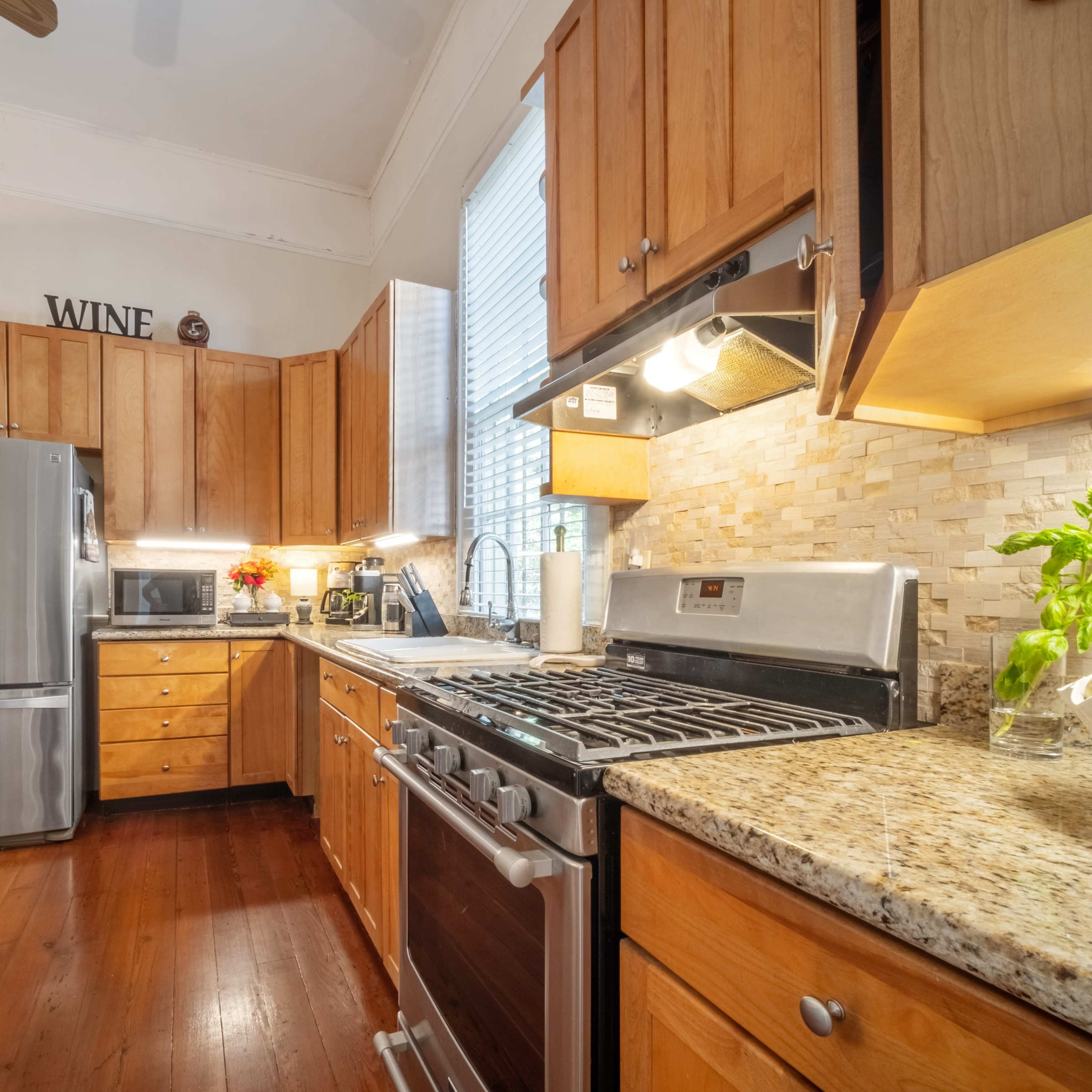 A modern kitchen features wooden cabinetry, stainless steel appliances, and granite countertops, with a view of a living area through an open doorway.