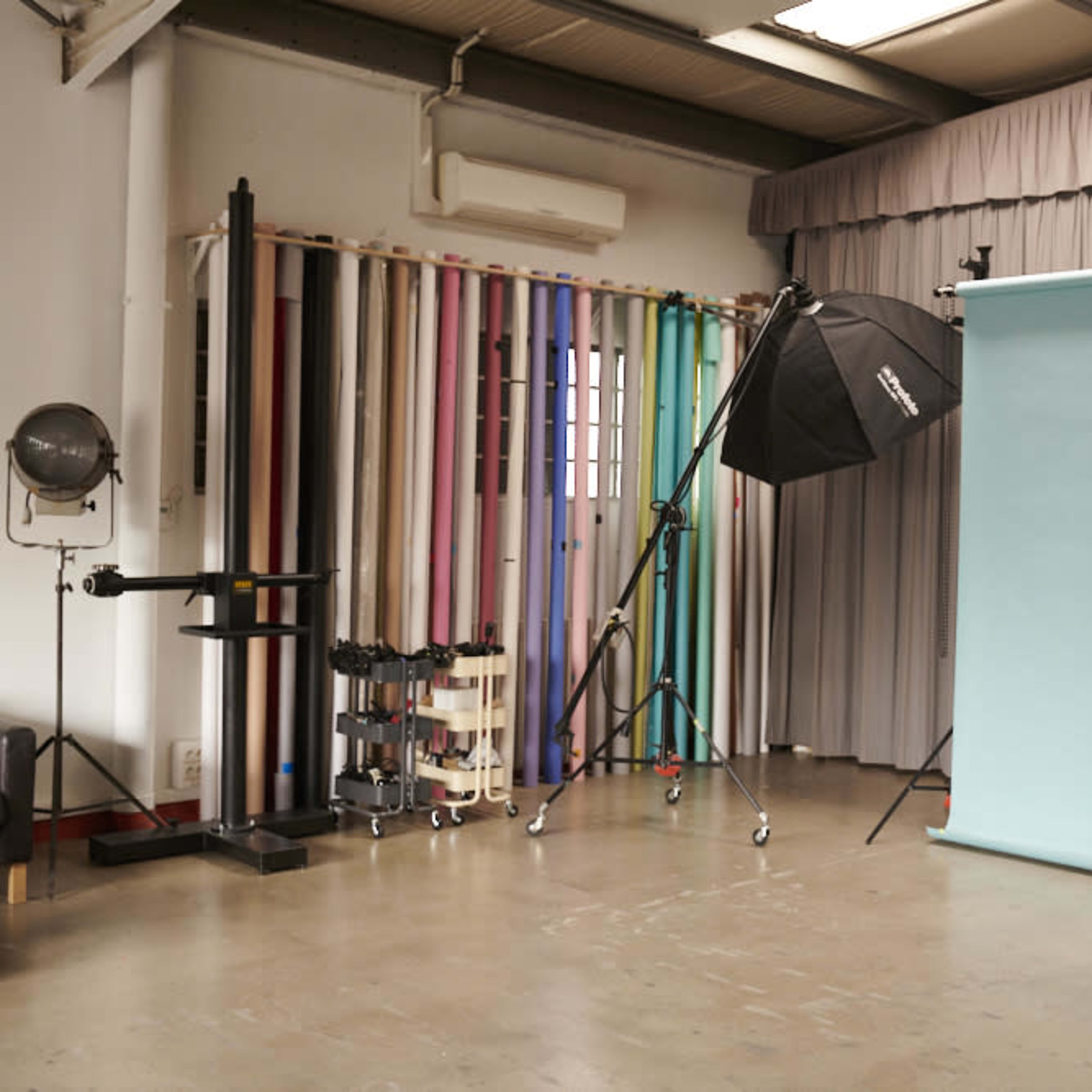 A photography studio features a light backdrop, lighting equipment, and colorful backdrop rolls arranged along a wall.