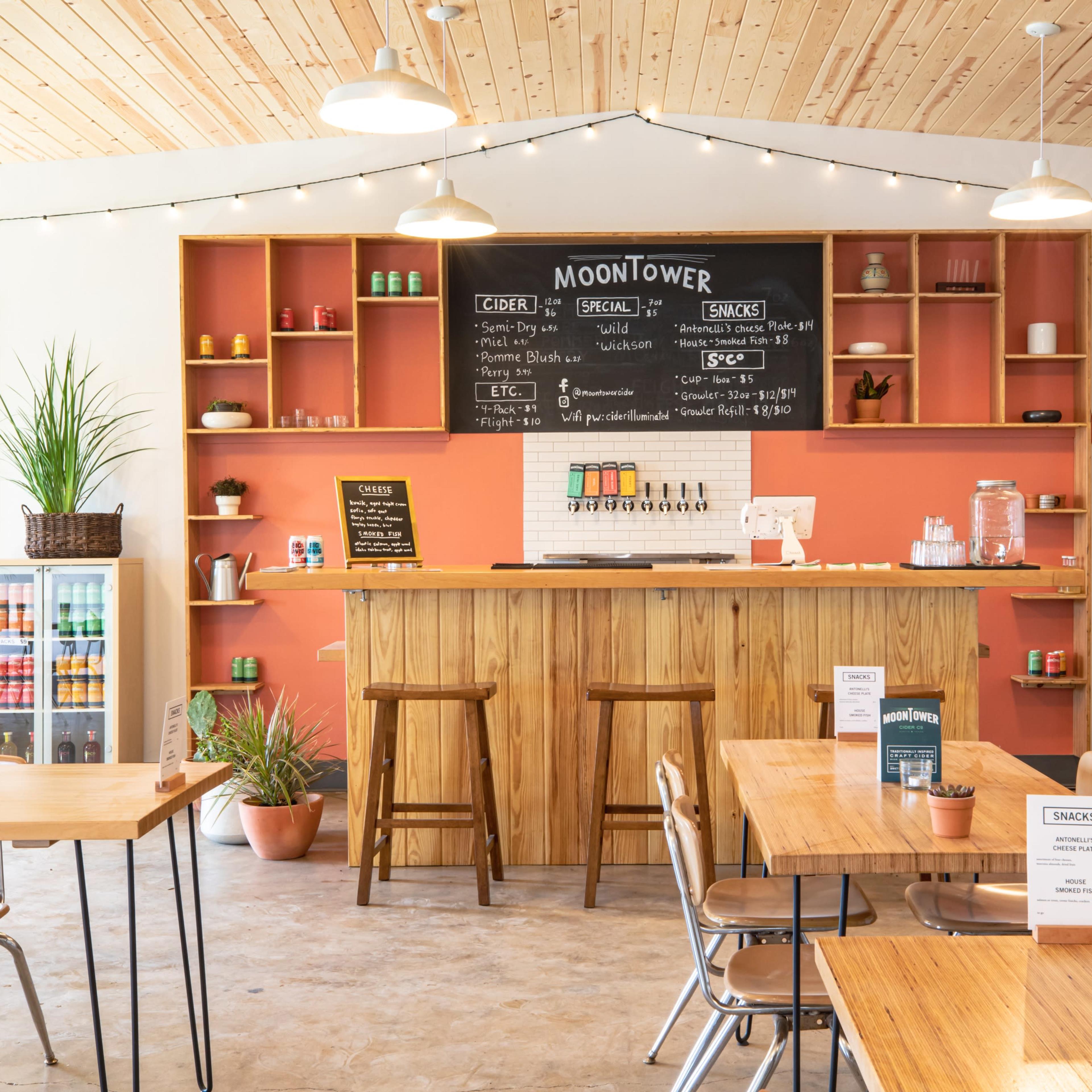 A cozy cider bar features a wooden counter, shelves with colorful bottles, and tables with menu signs under warm lighting.