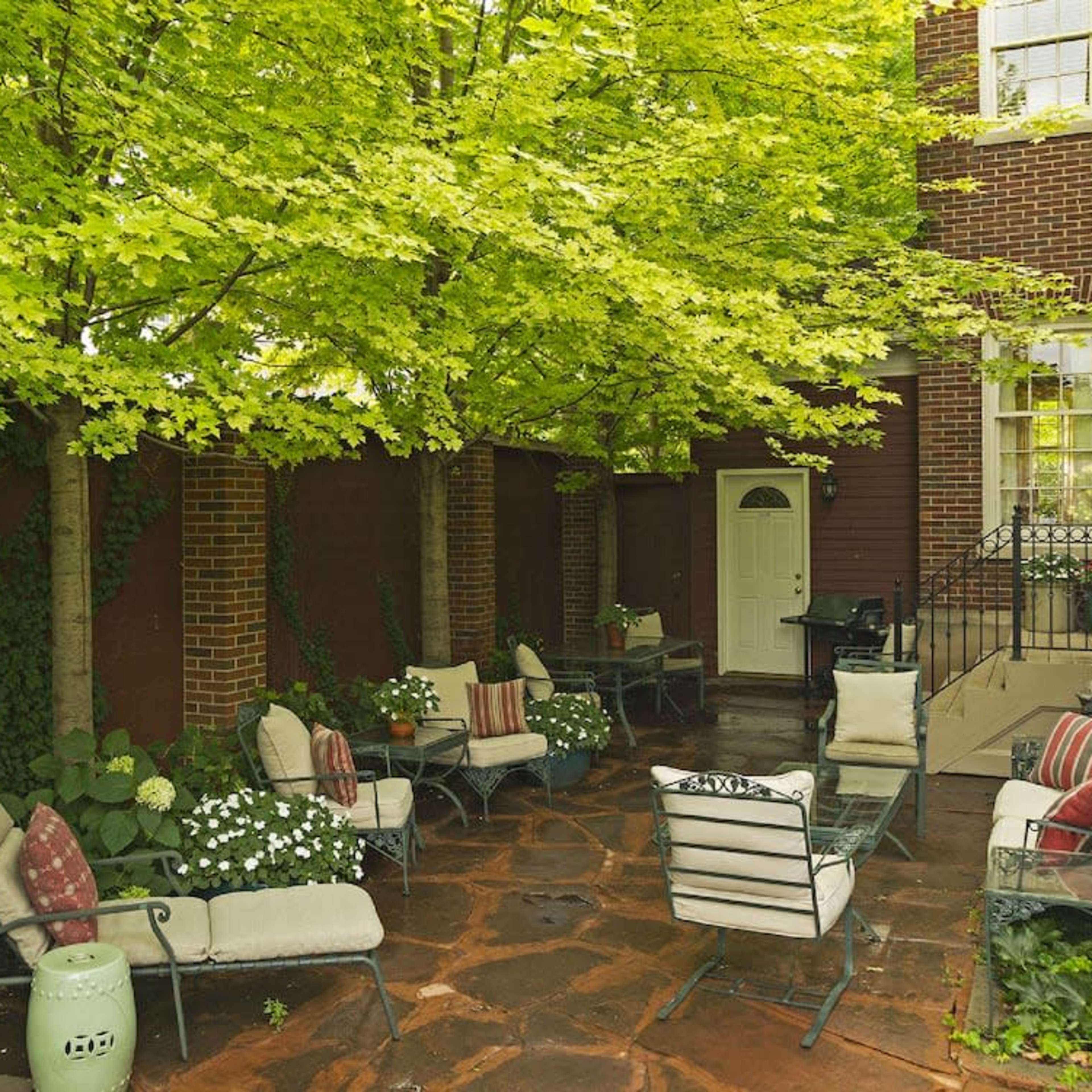 A patio area with stone flooring, surrounded by greenery, features several chairs and a door leading indoors.