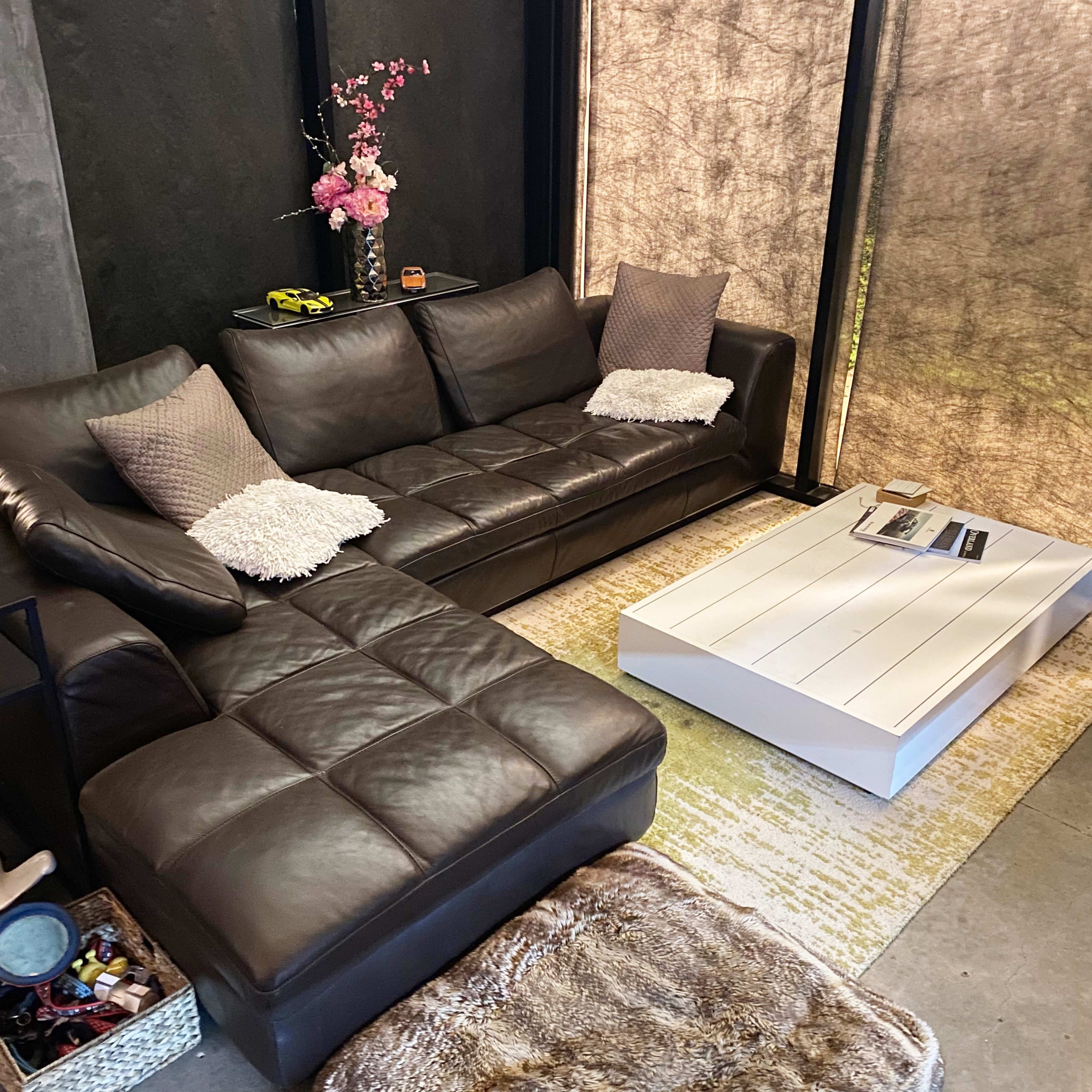 A modern living room featuring a dark leather sectional sofa, a white coffee table, and decorative accents on a gray wall with large windows covered by sheer curtains.