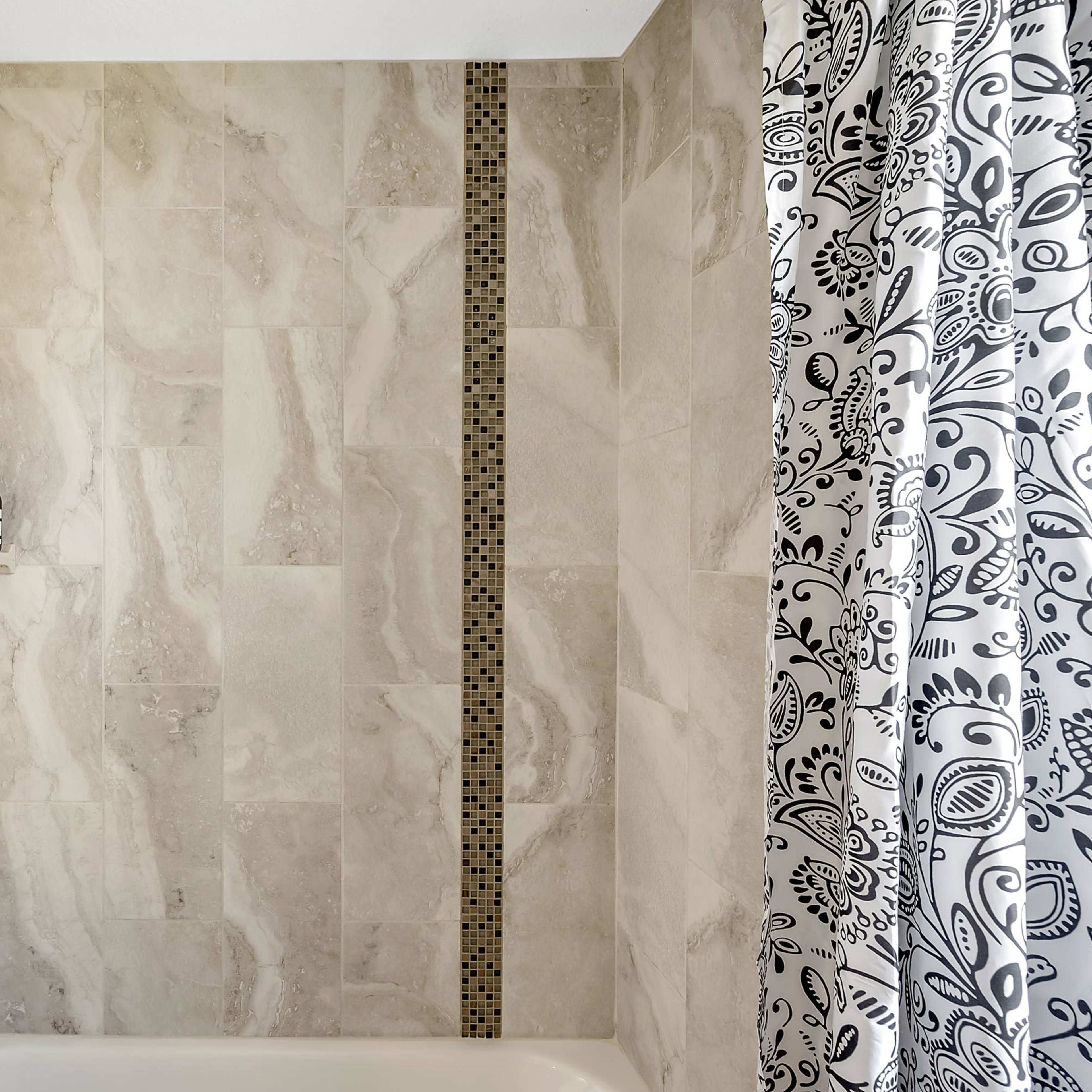 A shower area with beige tiled walls, a shower head, a shelf holding three bottles, and a patterned shower curtain.
