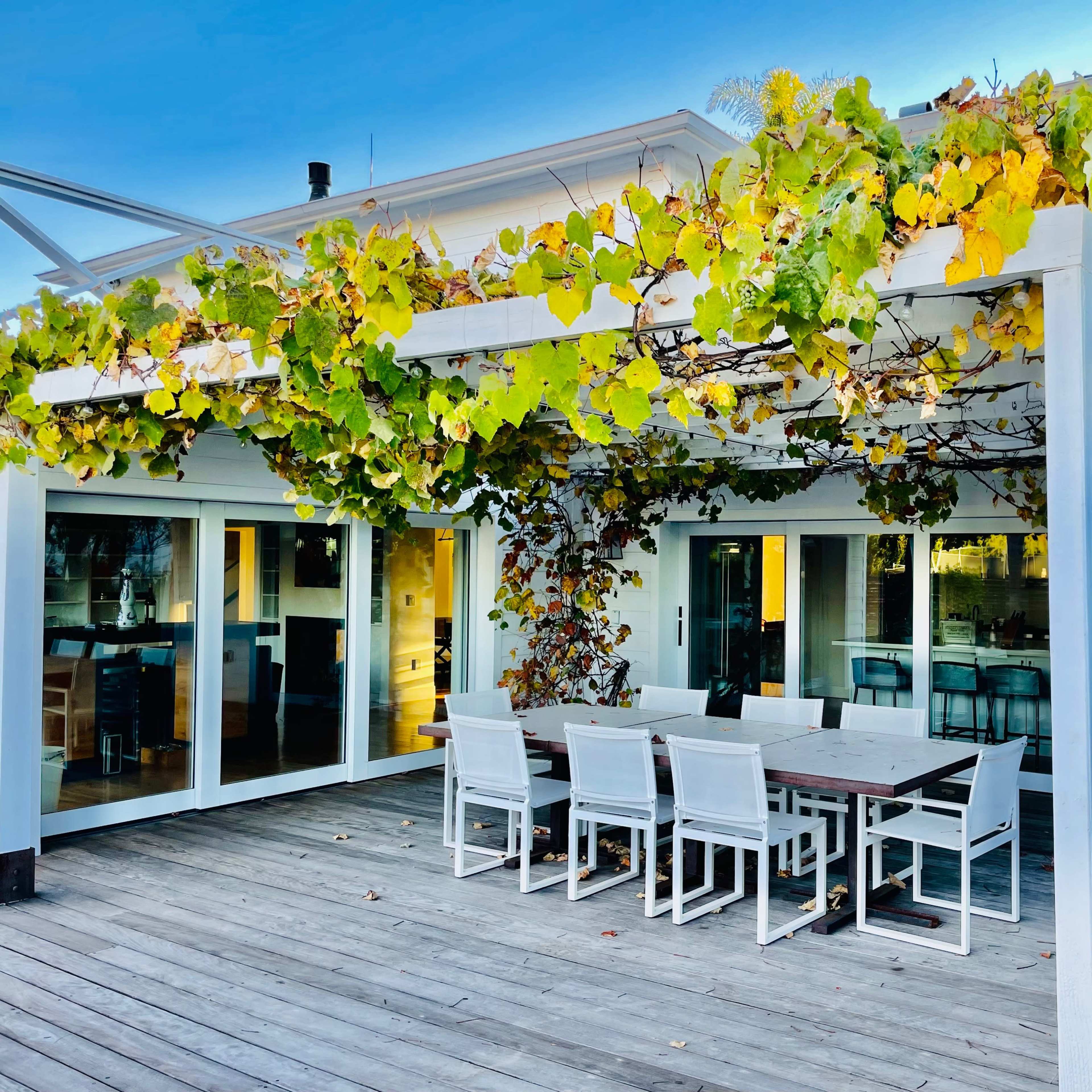 A patio with a dining table and chairs is shaded by a pergola covered in greenery, adjacent to a white house with large glass doors.