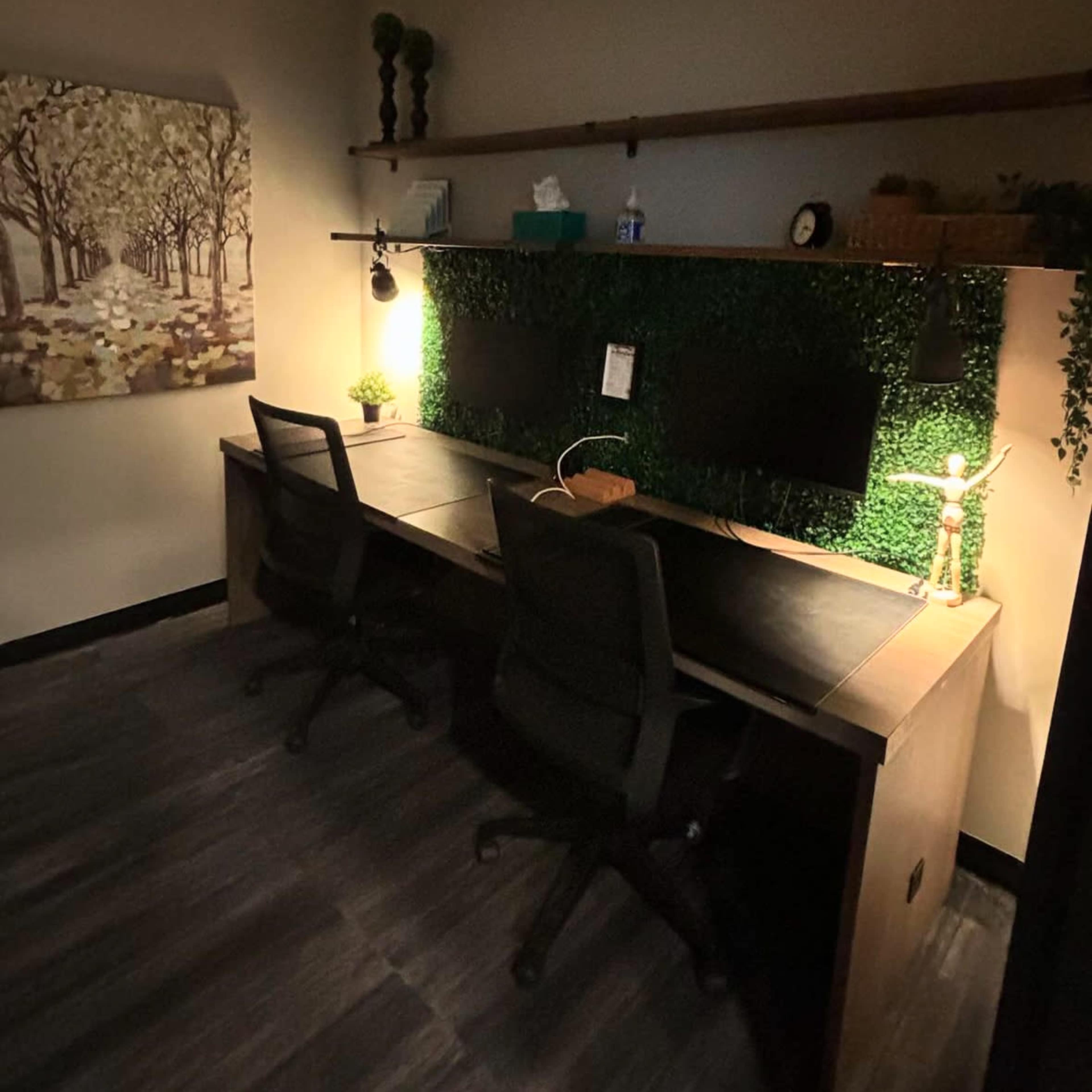 A dimly lit office space featuring a large desk with two black chairs, a wall adorned with faux greenery, and a monitor on the surface.
