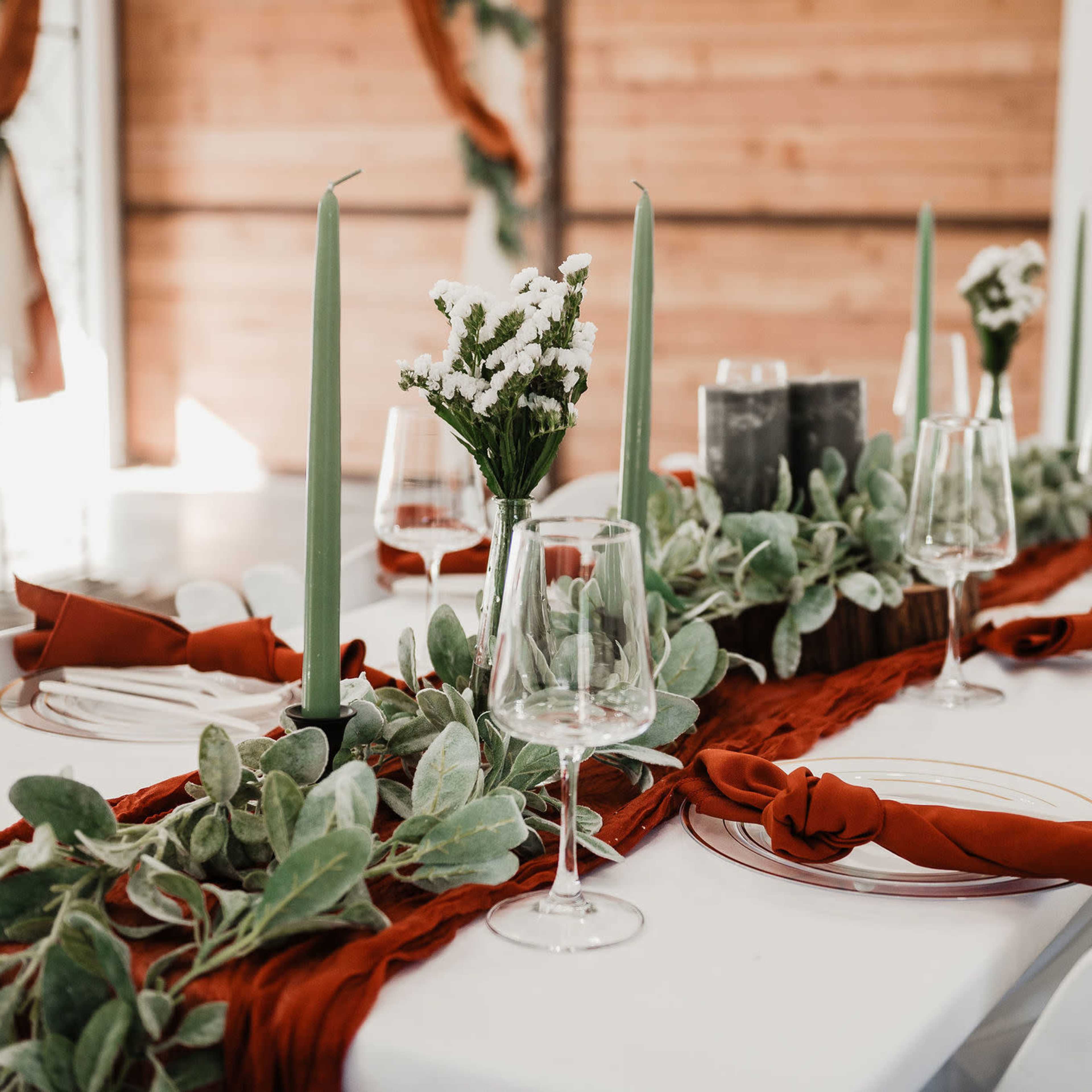 A dining table is set with green candles, floral arrangements, and a red cloth runner, accompanied by glassware and plates.