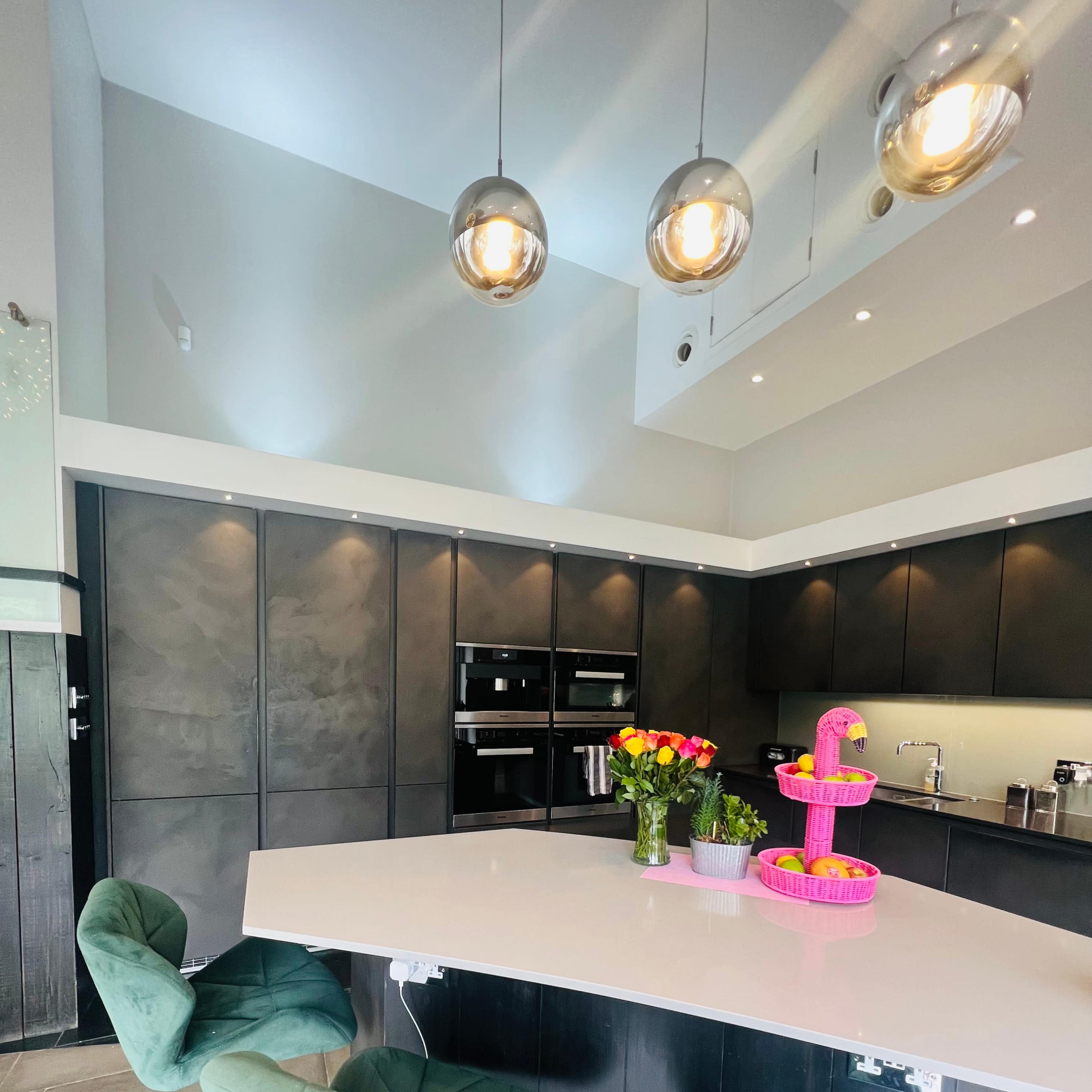 A modern kitchen features a circular island with green swivel chairs, overhead pendant lights, and a centerpiece of colorful flowers.
