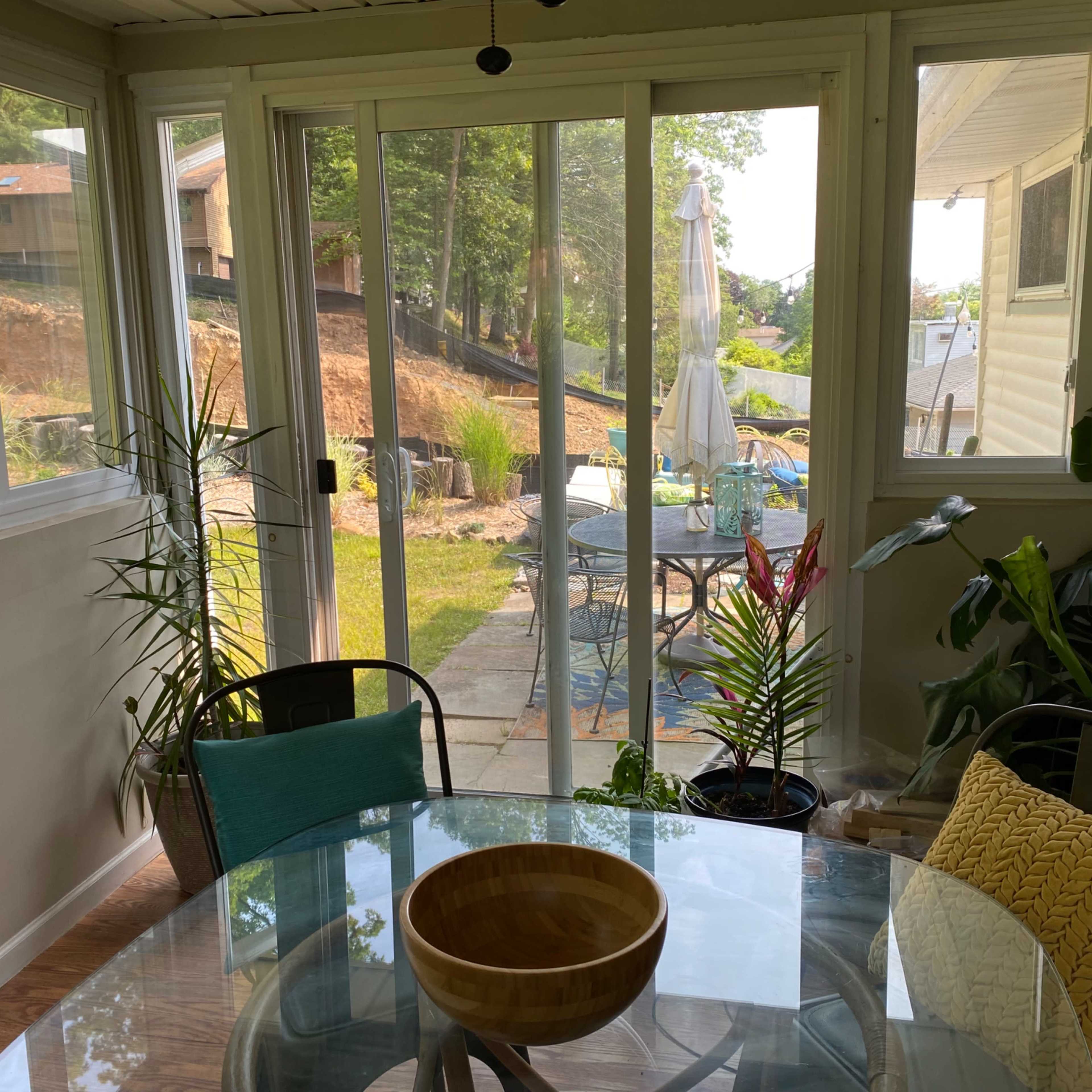 The scene features a dining area with a glass table and wooden bowl, looking through sliding doors to a patio and backyard with a table and chairs.