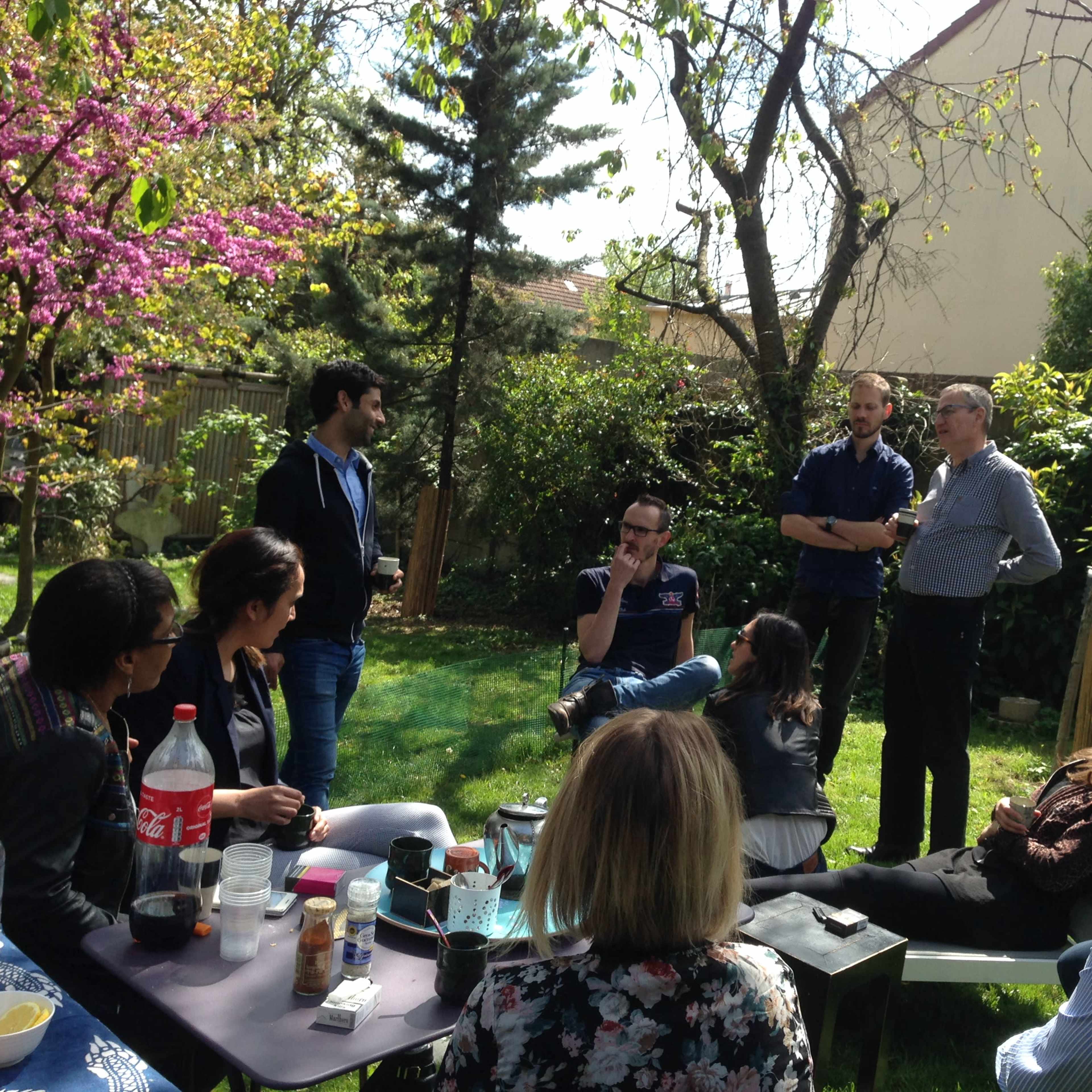 A group of people is gathered in a garden, engaging in conversation while seated and standing among greenery and blooming trees.