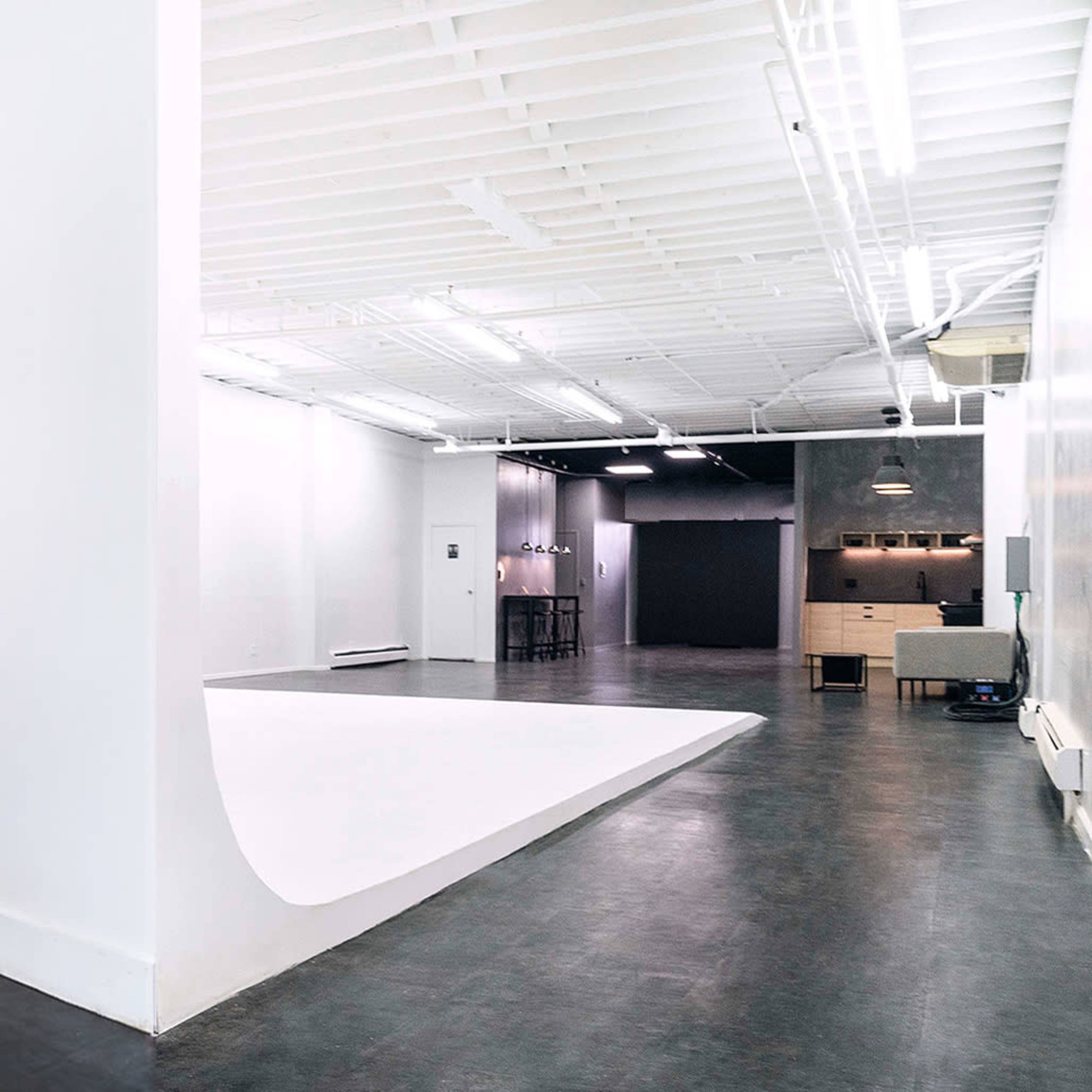 A spacious studio with white walls, a large white backdrop, and a minimalist design featuring a seating area and a small kitchenette.