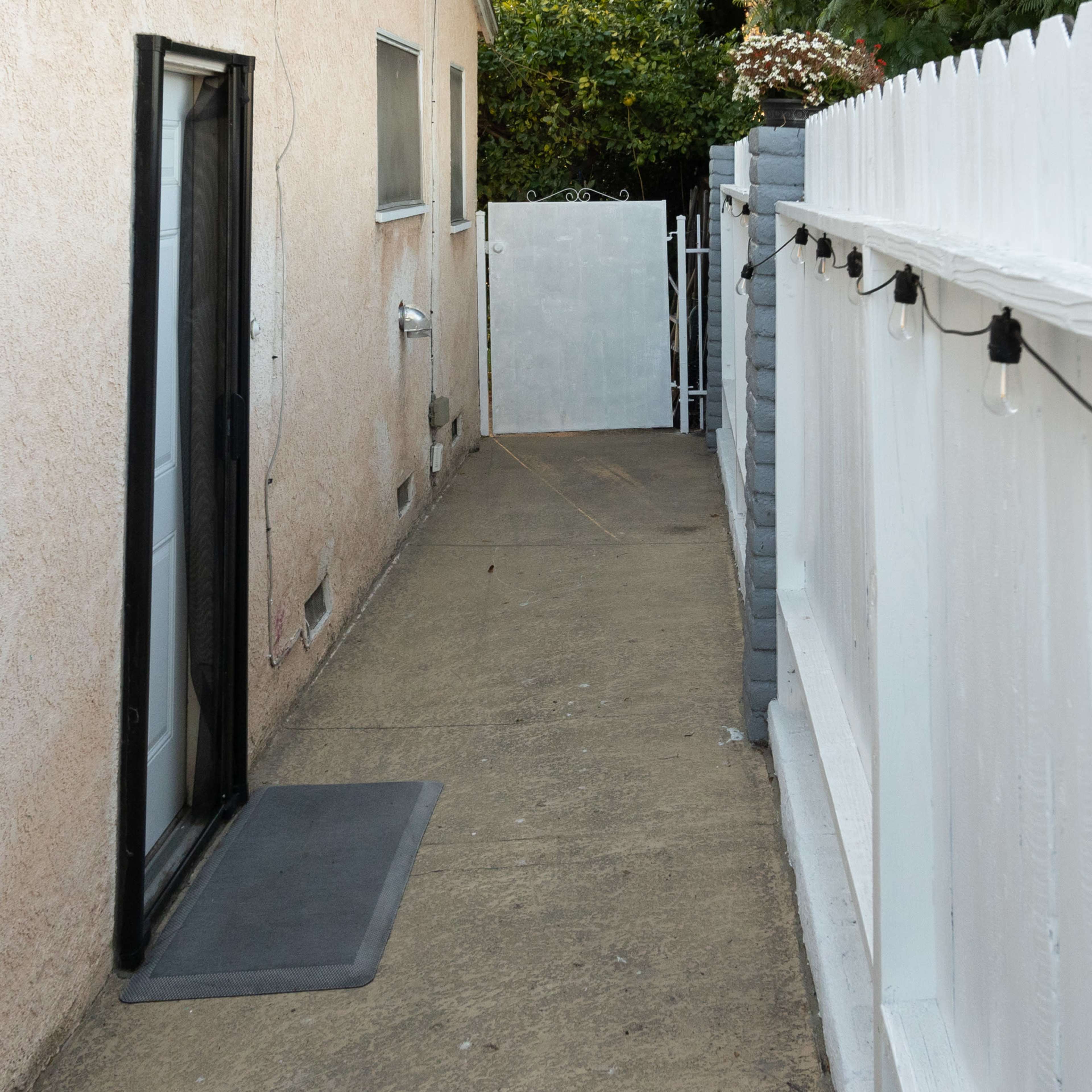 A narrow concrete walkway runs alongside a house, bordered by a white fence and a door at the far end.