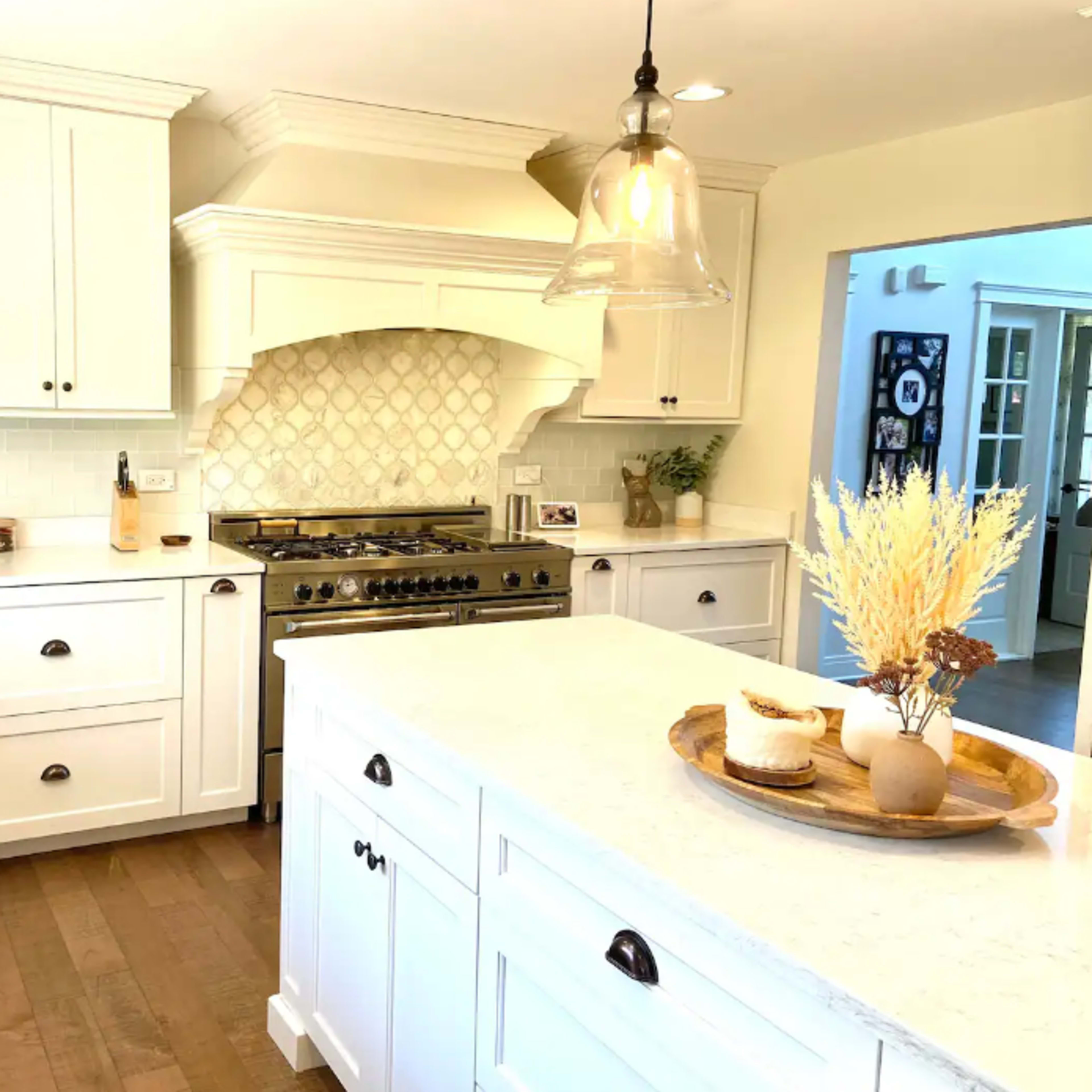 A modern kitchen featuring white cabinetry, a large island with a wooden tray, and a gas range with a decorative hood.
