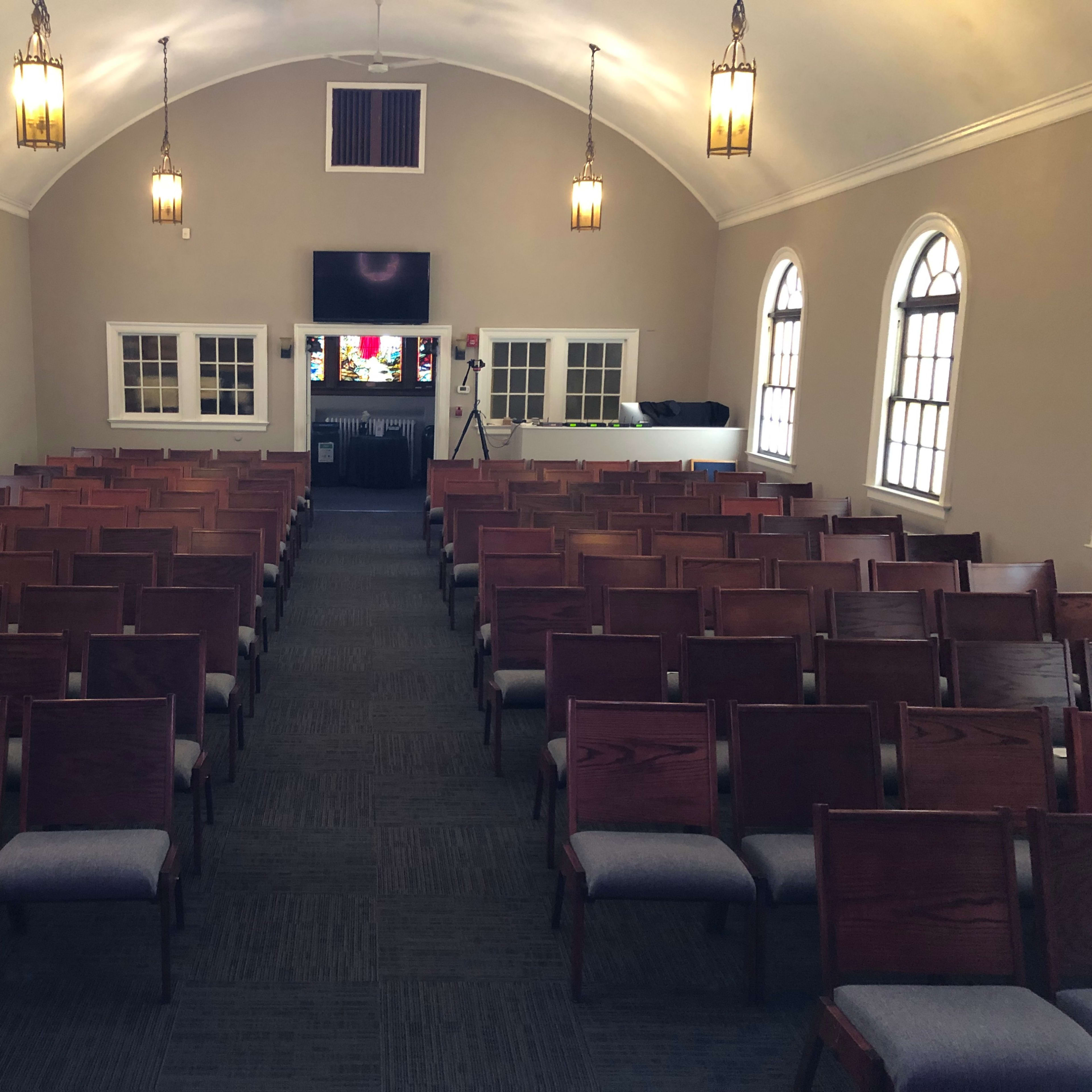 The interior of a church with rows of wooden chairs facing a stage and large windows on either side.