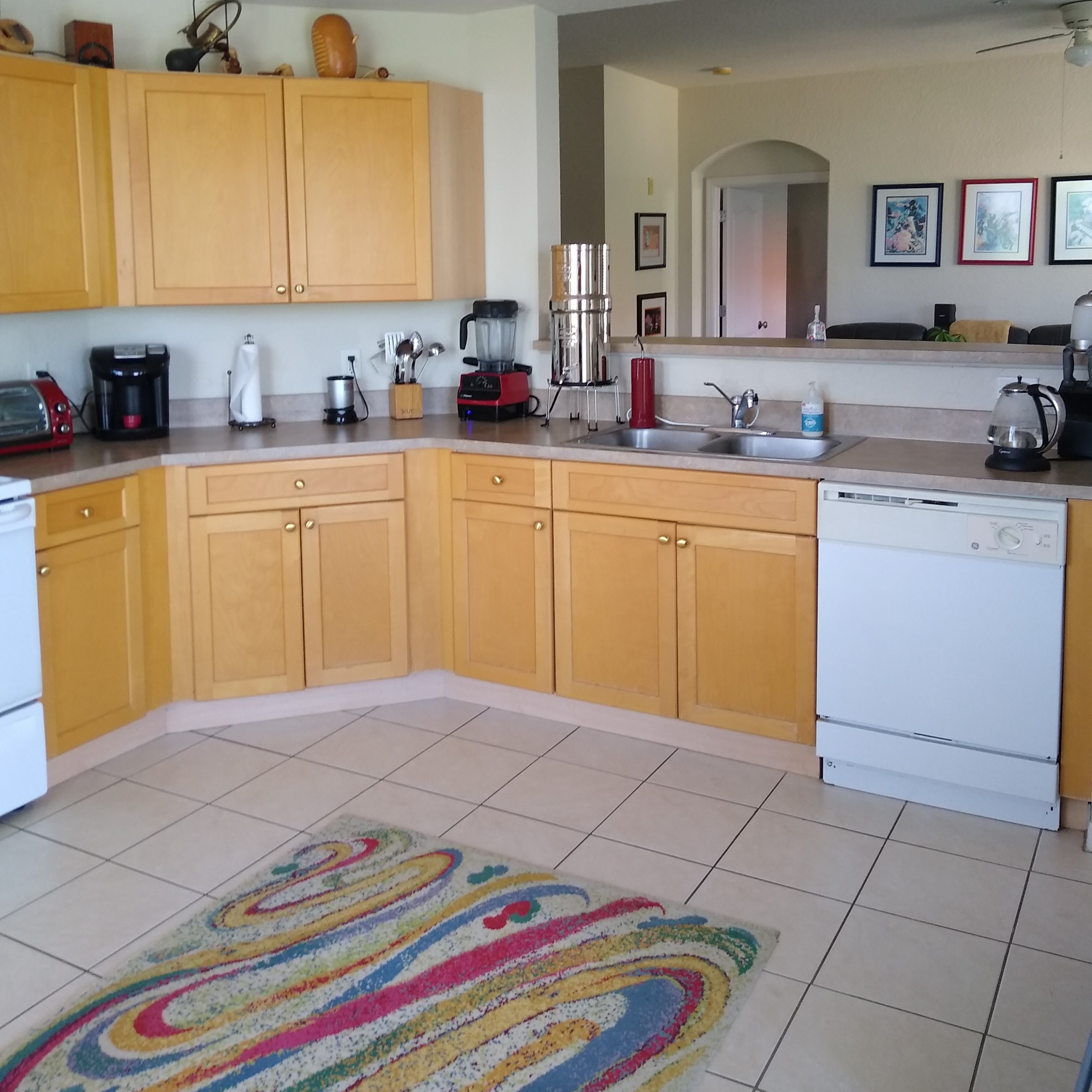A spacious kitchen with yellow wooden cabinets, a countertop with appliances, and tiled flooring featuring a colorful rug.
