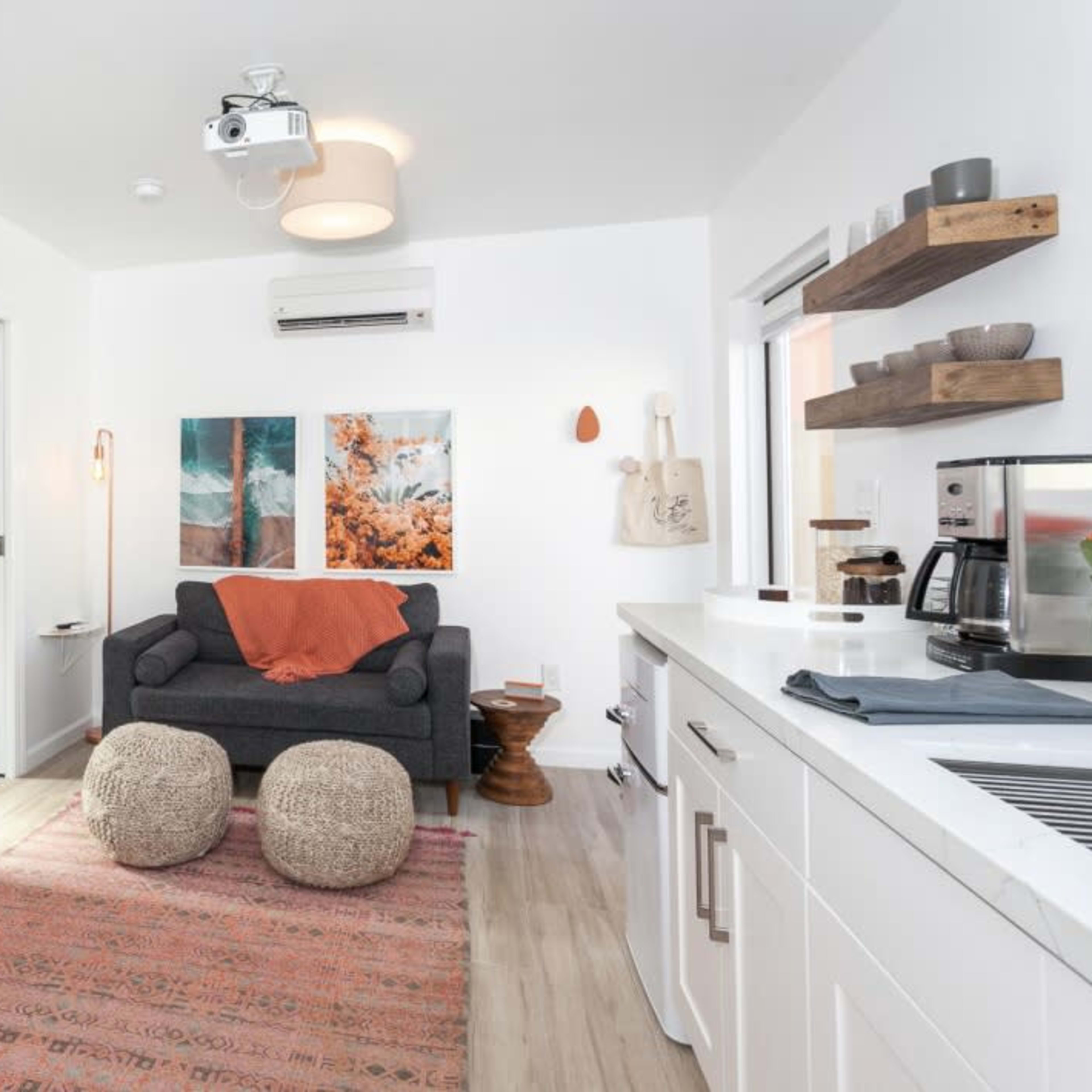 A small living space features a gray couch with an orange throw blanket, a patterned rug, two art pieces on the wall, and a kitchen area with a coffee maker and open shelving.