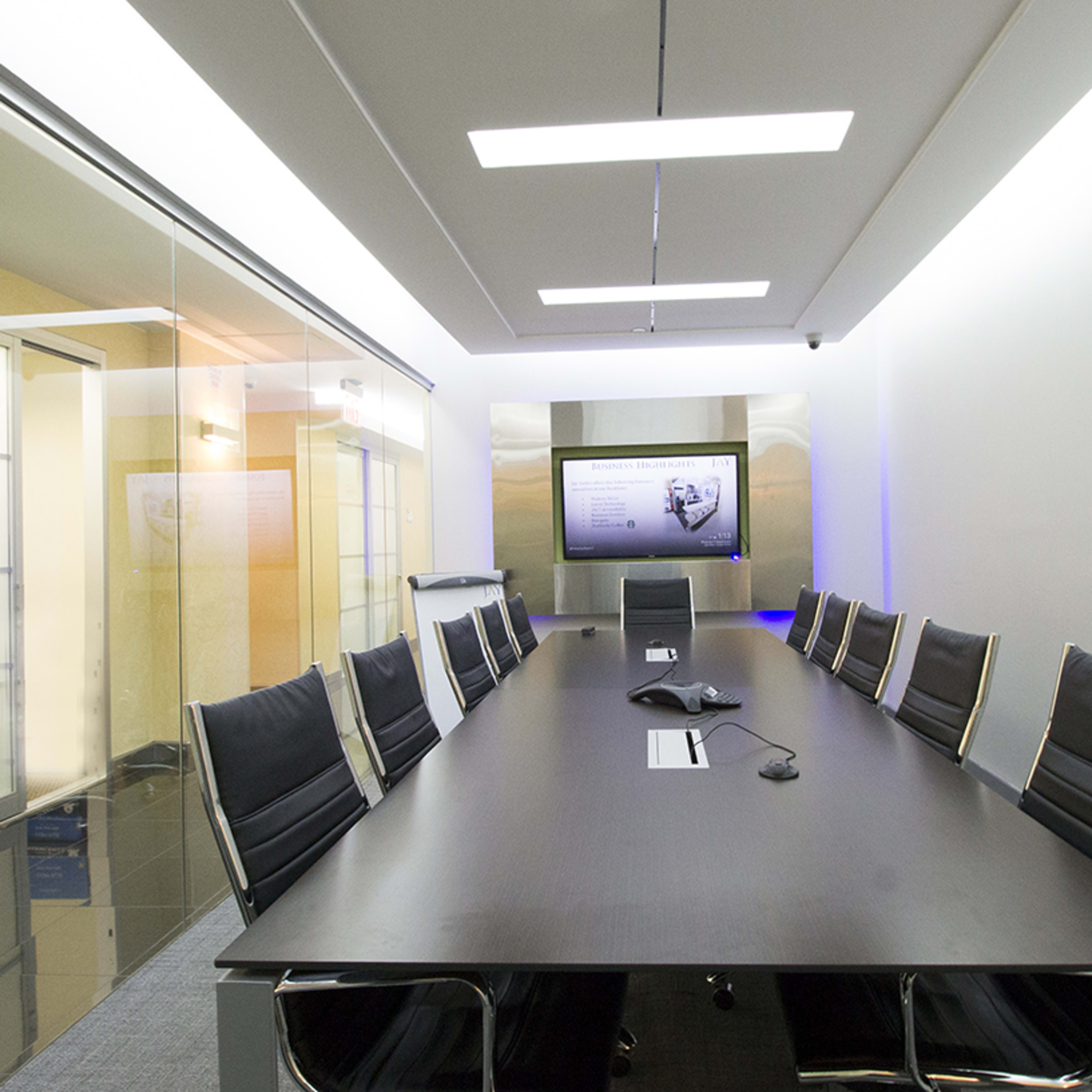 A modern conference room features a long table surrounded by black chairs, with a large screen displaying a presentation on the wall.