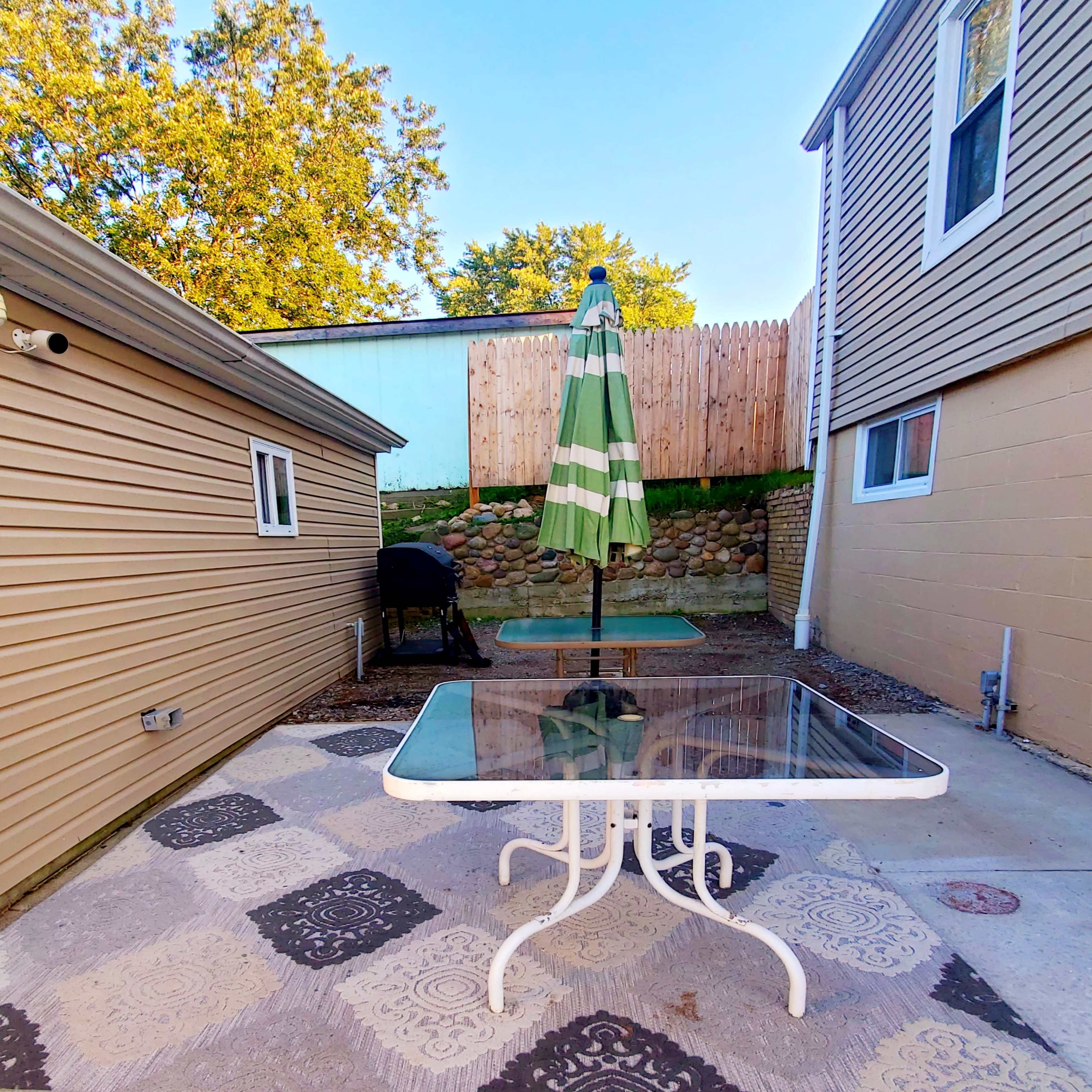 A patio area features a glass-top table with a green and white umbrella surrounded by two buildings and a stone wall.